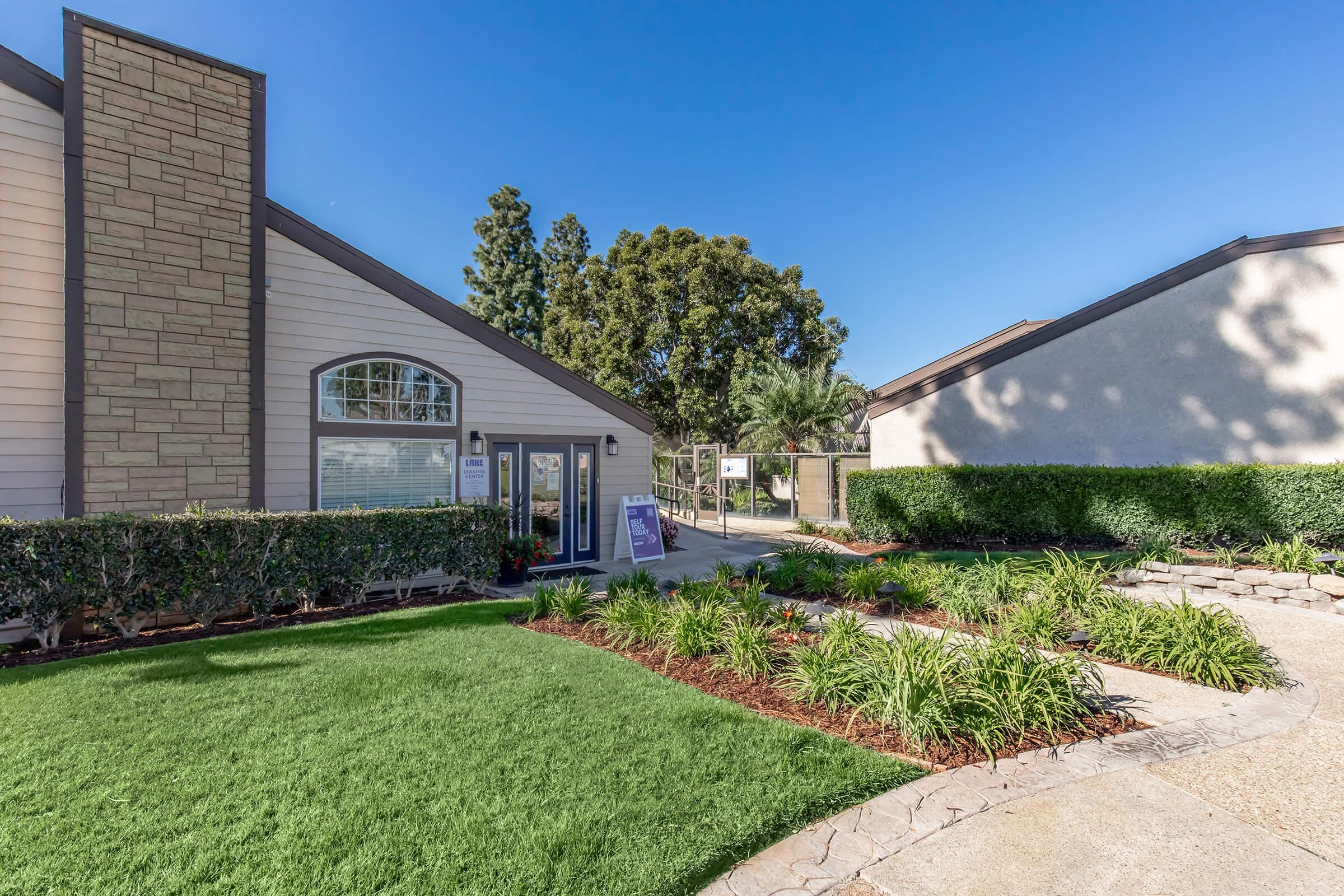 A well-maintained exterior of a building with a stone façade, surrounded by lush greenery and landscaped gardens. The pathway leads up to the entrance, which has large windows and a welcoming atmosphere. Blue skies and trees are visible in the background, enhancing the serene setting.
