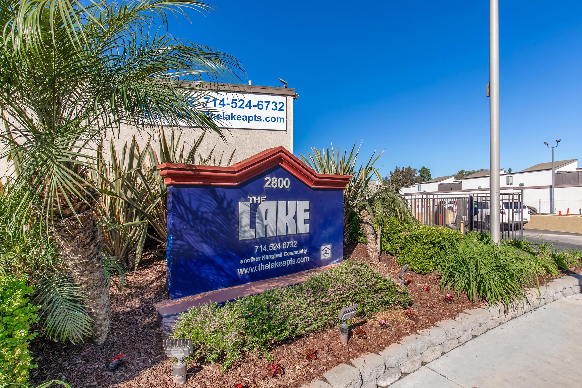 Sign for "The Lake" apartments at 2800, featuring contact number 714-524-6732. The sign is surrounded by palm trees and colorful landscaping, with a clear blue sky in the background.