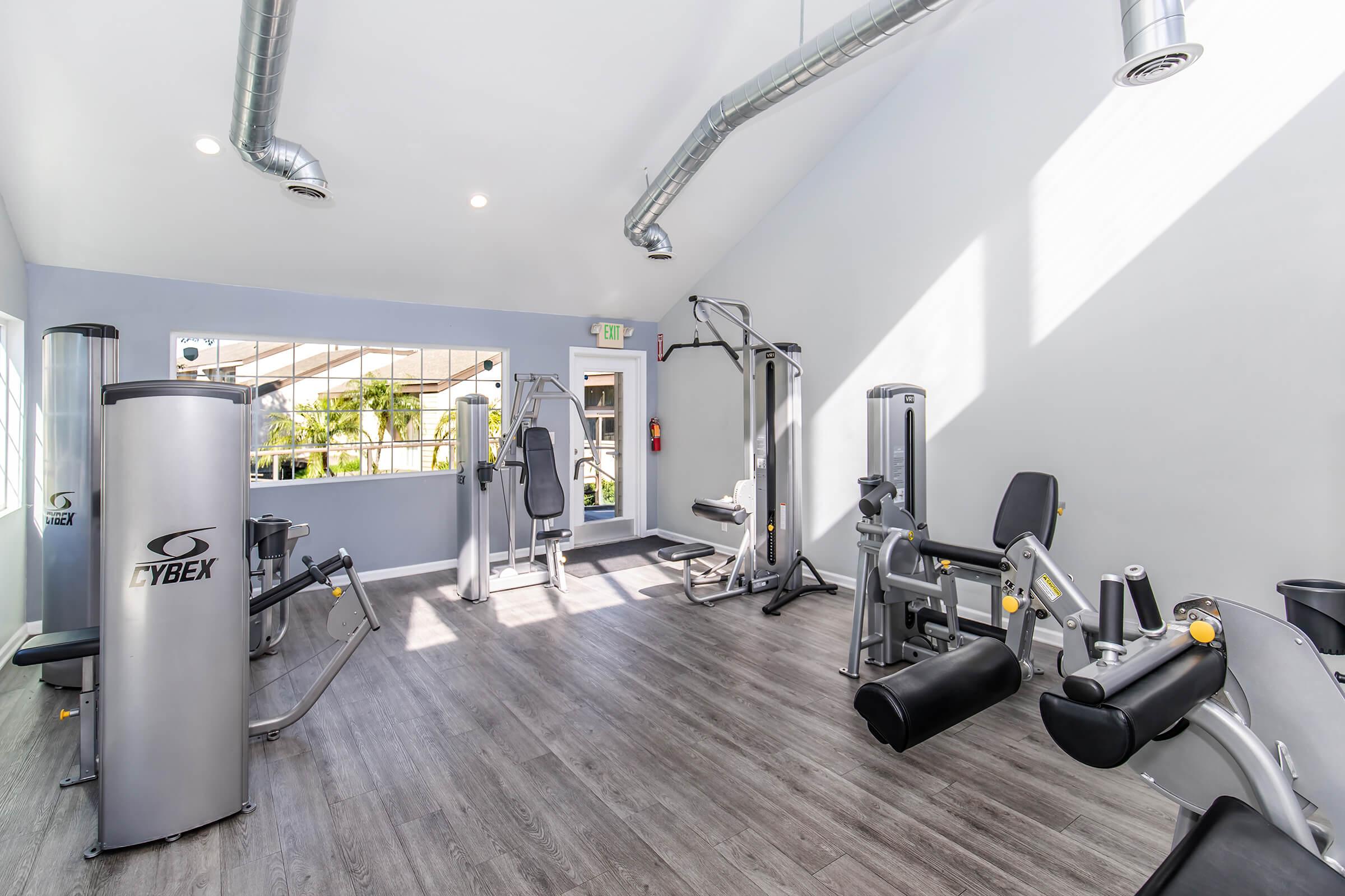 A well-lit gym interior featuring modern exercise machines including a leg press, weight machines, and cardio equipment. The space has large windows, a gray color scheme, and wooden flooring, creating a clean and inviting atmosphere for workouts.