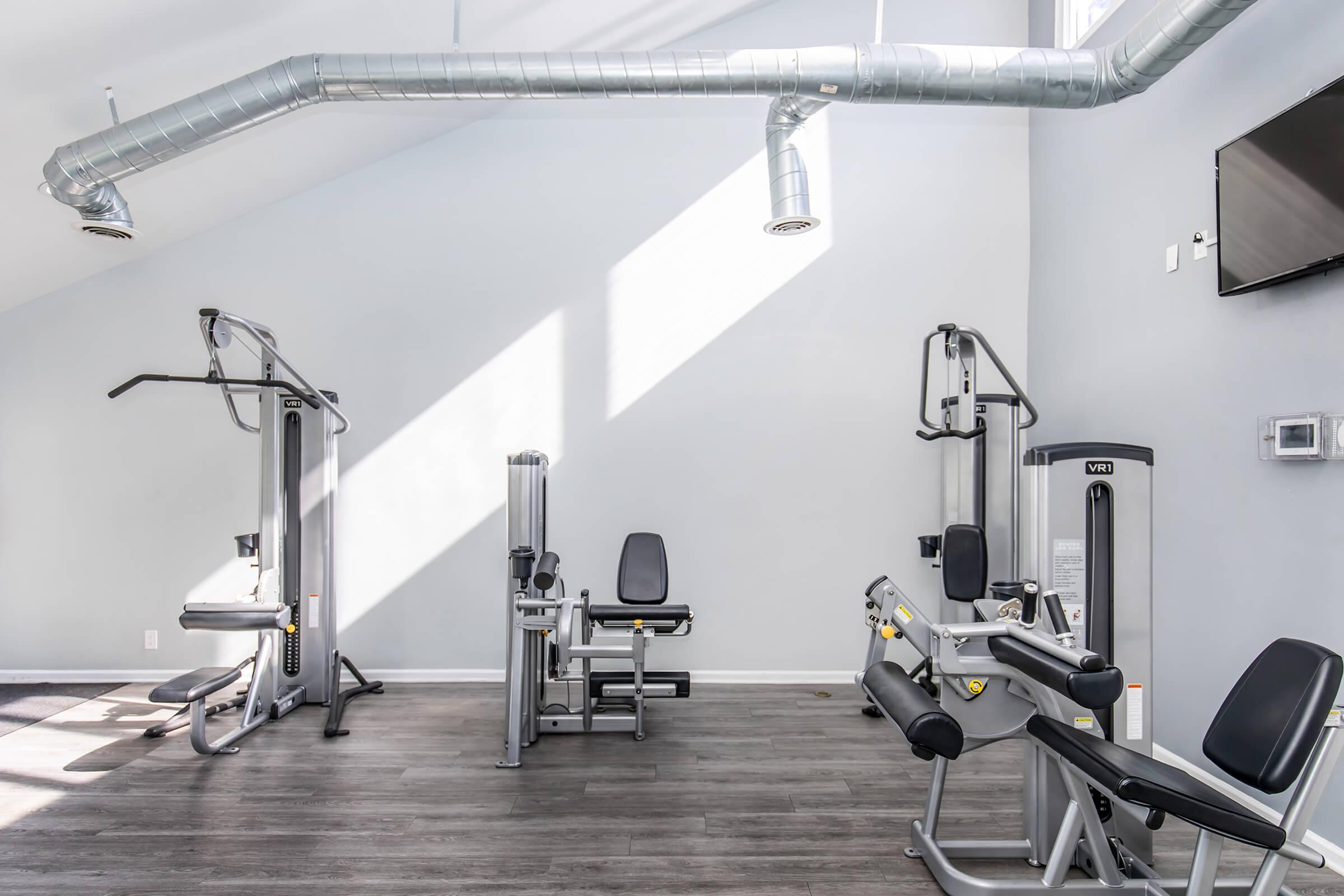 A bright, modern gym with gray walls and large windows. Fitness equipment includes a cable machine, leg press, and multi-use exercise machine. The flooring is wood-like, and there is a flat-screen TV mounted on the wall, enhancing the space. Natural light streams in, creating an inviting atmosphere for workouts.