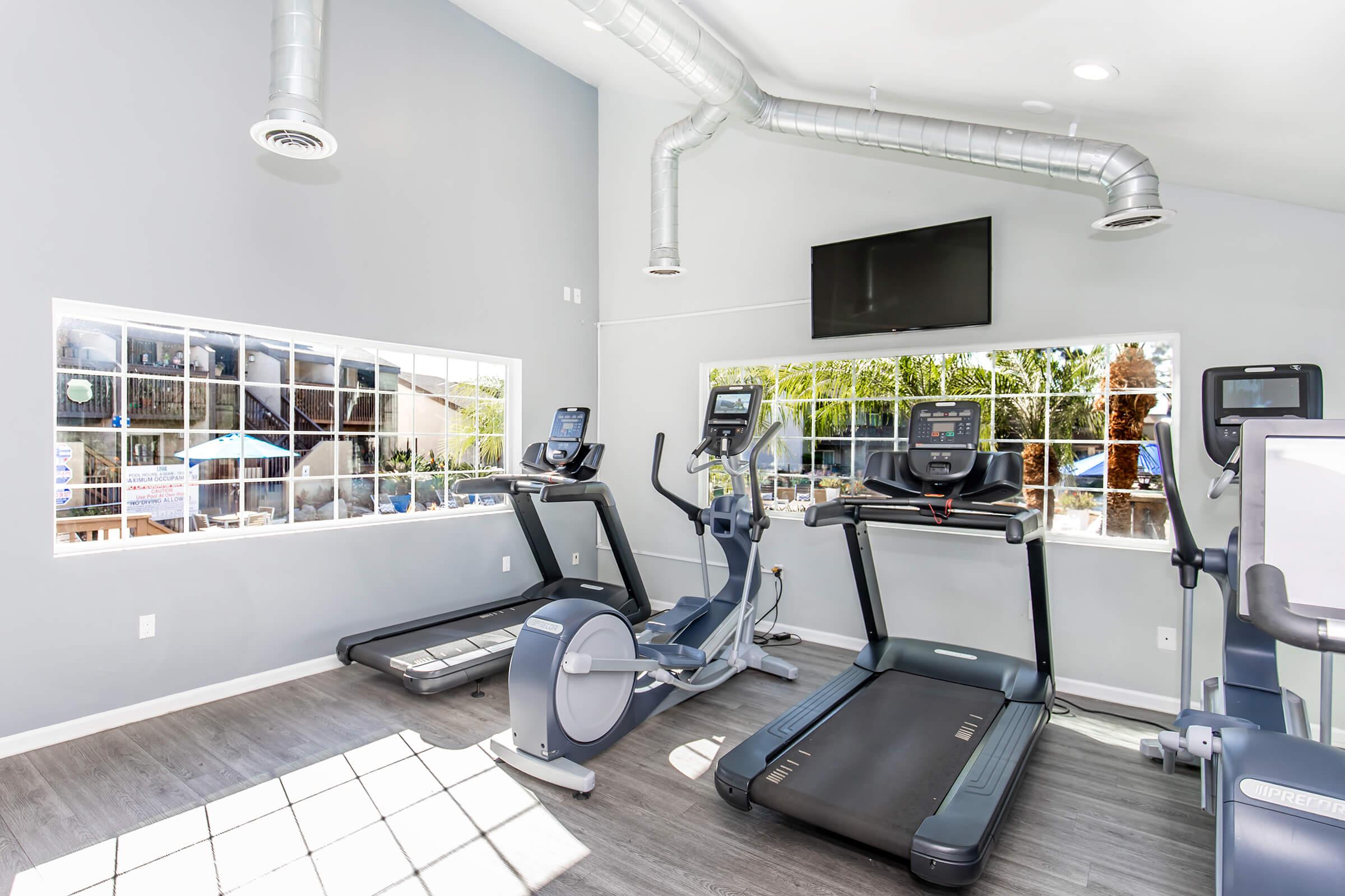 A modern gym featuring a treadmill, elliptical machine, and stationary bike. Large windows allow natural light to illuminate the space, which has a neutral color scheme and sleek equipment. A mounted television is visible on the wall, enhancing the workout environment.