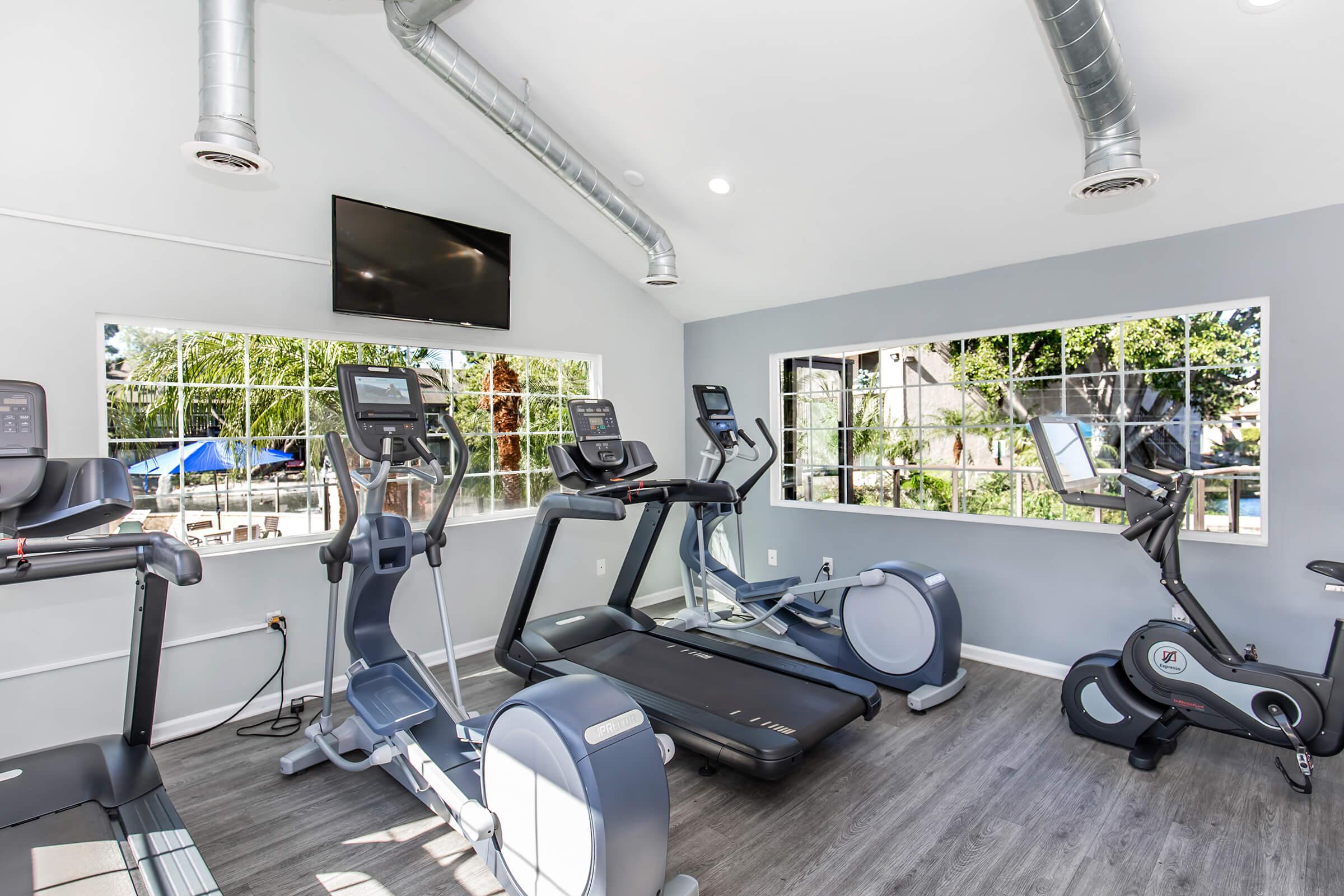 A modern gym interior featuring various exercise equipment, including treadmills and stationary bikes. Large windows allow natural light to fill the space, providing views of an outdoor pool area. A wall-mounted television is visible. The decor is sleek with gray walls and wood-like flooring.