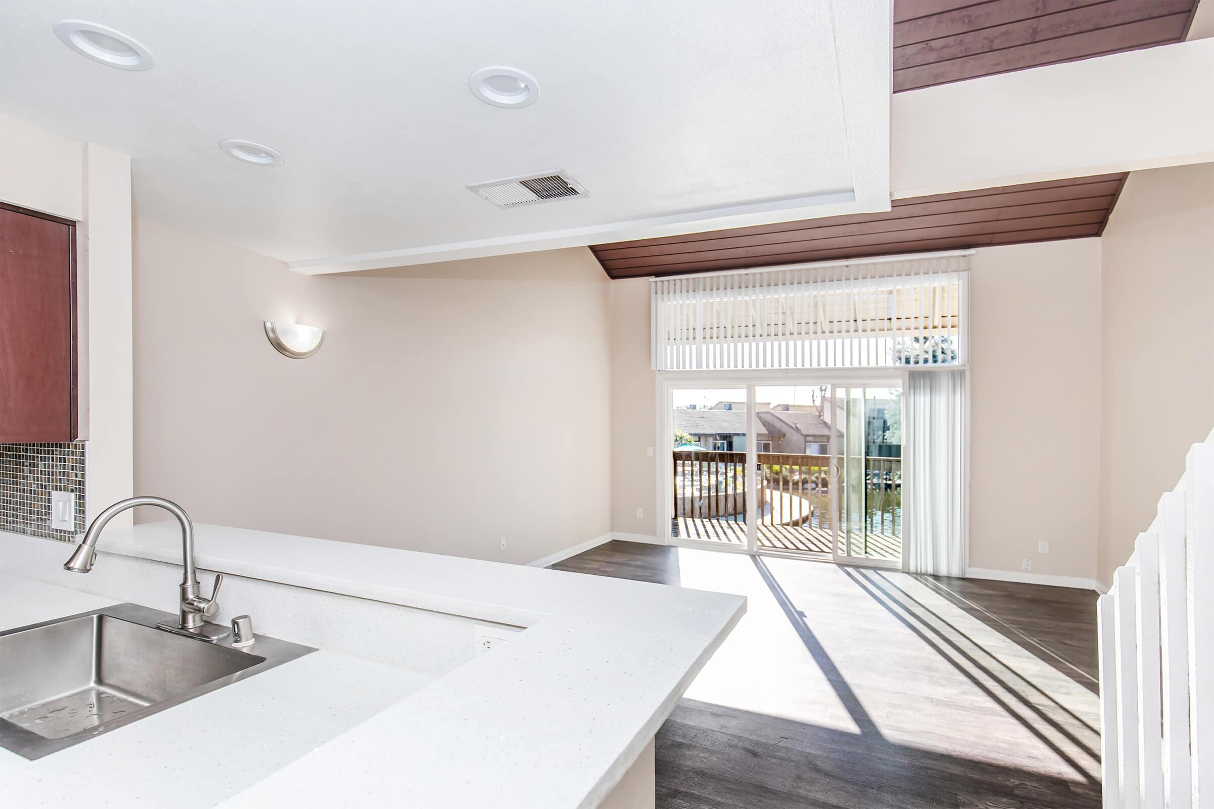 Modern open-concept interior featuring a kitchen island with a sink, overlooking a spacious living area with large windows and sliding glass doors that lead to a balcony. The walls are painted in light neutral tones, complemented by wood accents on the ceiling and floor, creating a bright and inviting atmosphere.