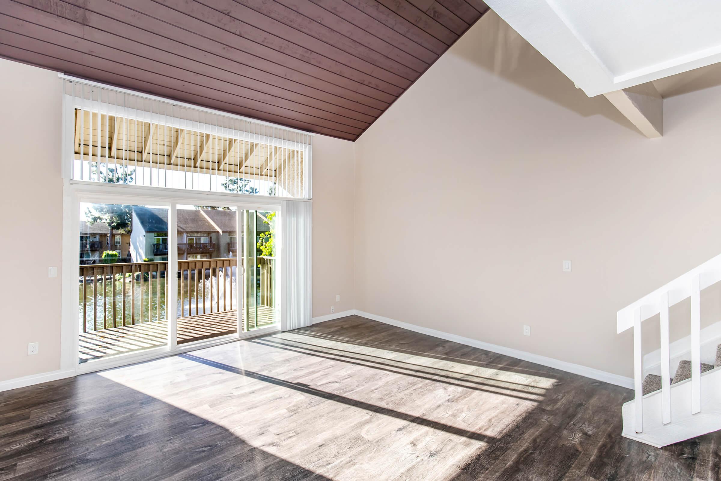 A bright, spacious living room with large windows overlooking a deck by a waterway. The room features high ceilings with wooden beams, light-colored walls, and new flooring. Natural light streams in, creating an open and inviting atmosphere. A staircase is visible in the corner, leading to an upper level.
