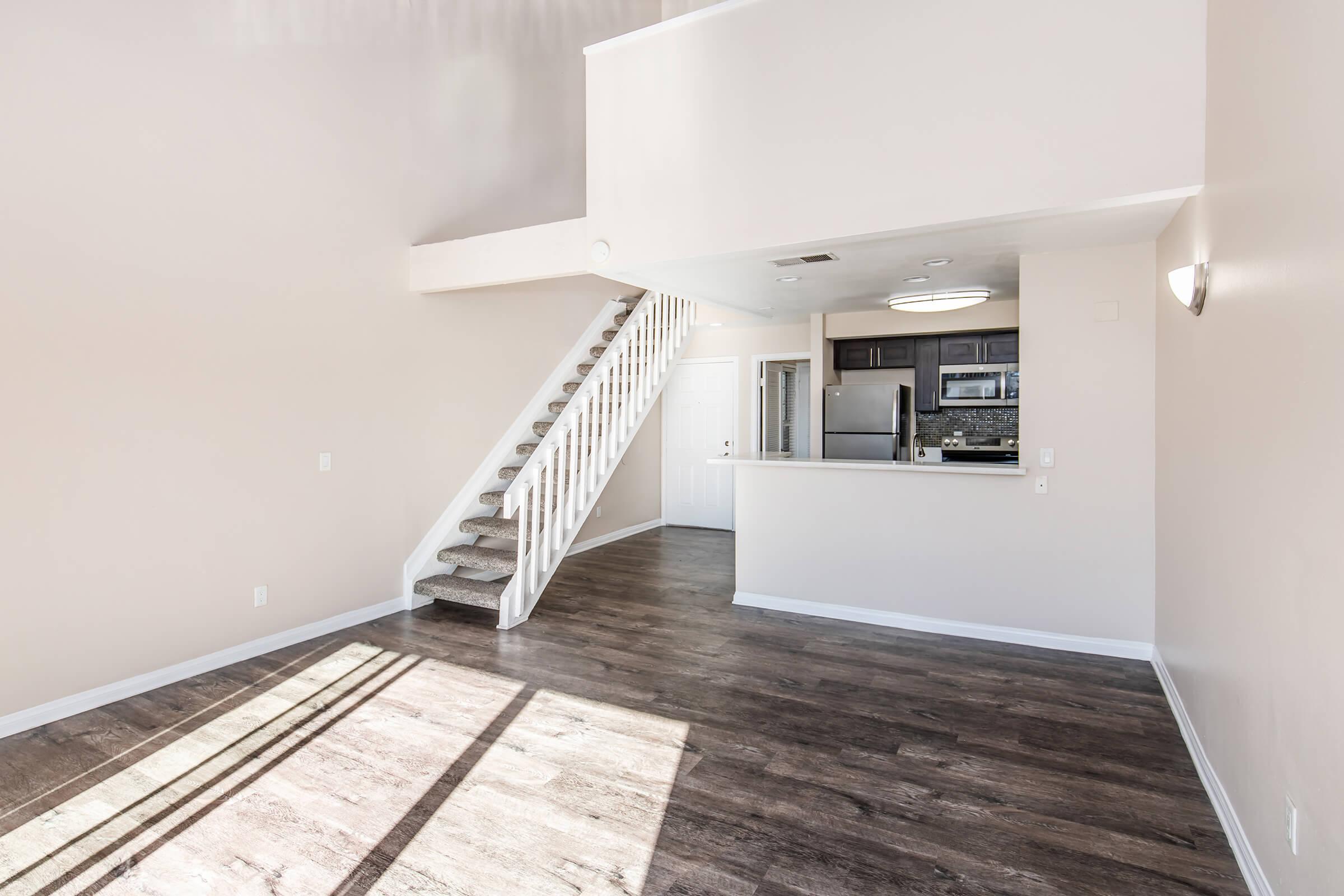 A well-lit, modern interior featuring a living area with wood-style flooring, a staircase leading to a second level, and an open kitchen with dark cabinetry and stainless steel appliances. The walls are painted a soft beige, and natural light streams in through a window.