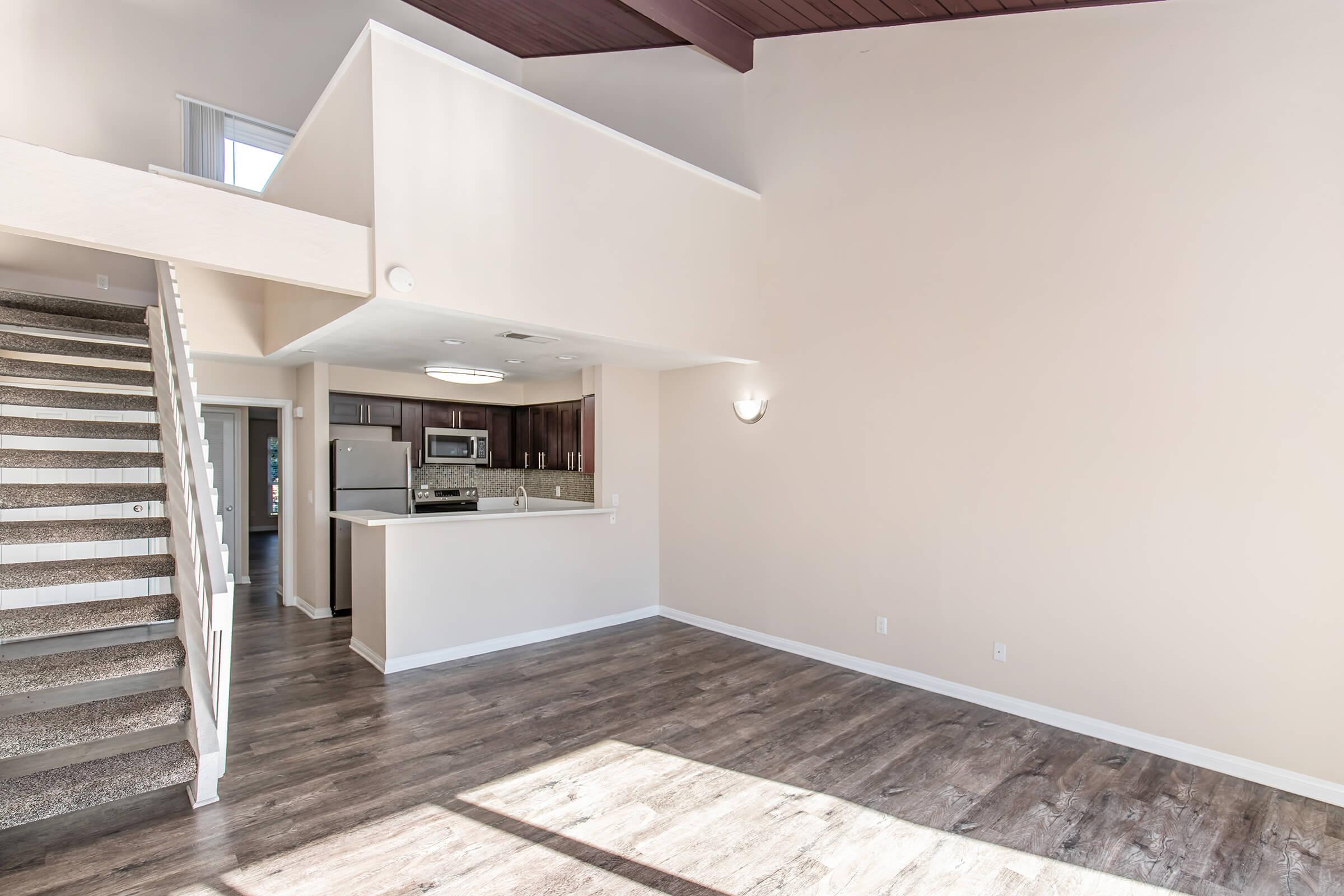 Spacious interior of a modern living area featuring a two-story design. It includes a staircase with a textured railing, a kitchen with dark cabinetry, stainless steel appliances, and a light-colored wall. Large windows allow natural light to fill the room, creating an inviting atmosphere.