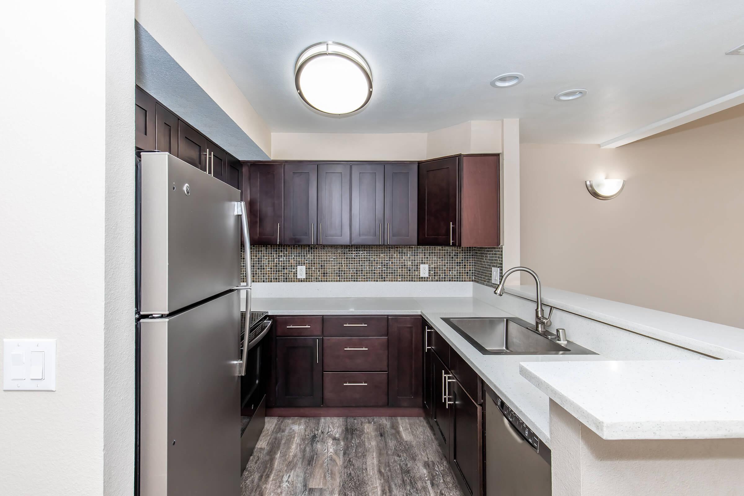 A modern kitchen featuring dark cabinetry, stainless steel appliances, and a sleek countertop. The kitchen includes a double sink, a mosaic backsplash, and an open layout that connects to the living area. Soft lighting enhances the contemporary design.