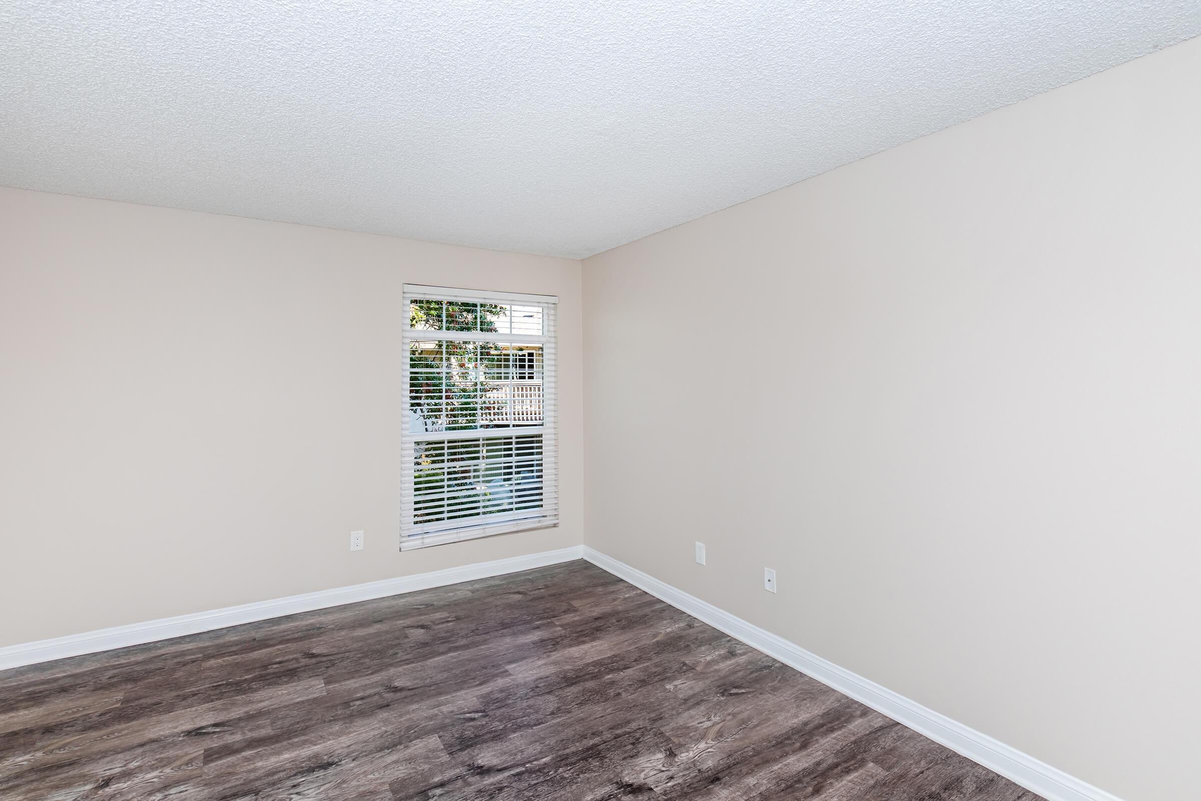 Empty room with light beige walls and a wood-like floor. A small window with horizontal blinds allows natural light to enter. The space appears clean and uncluttered, suitable for various uses or decoration.