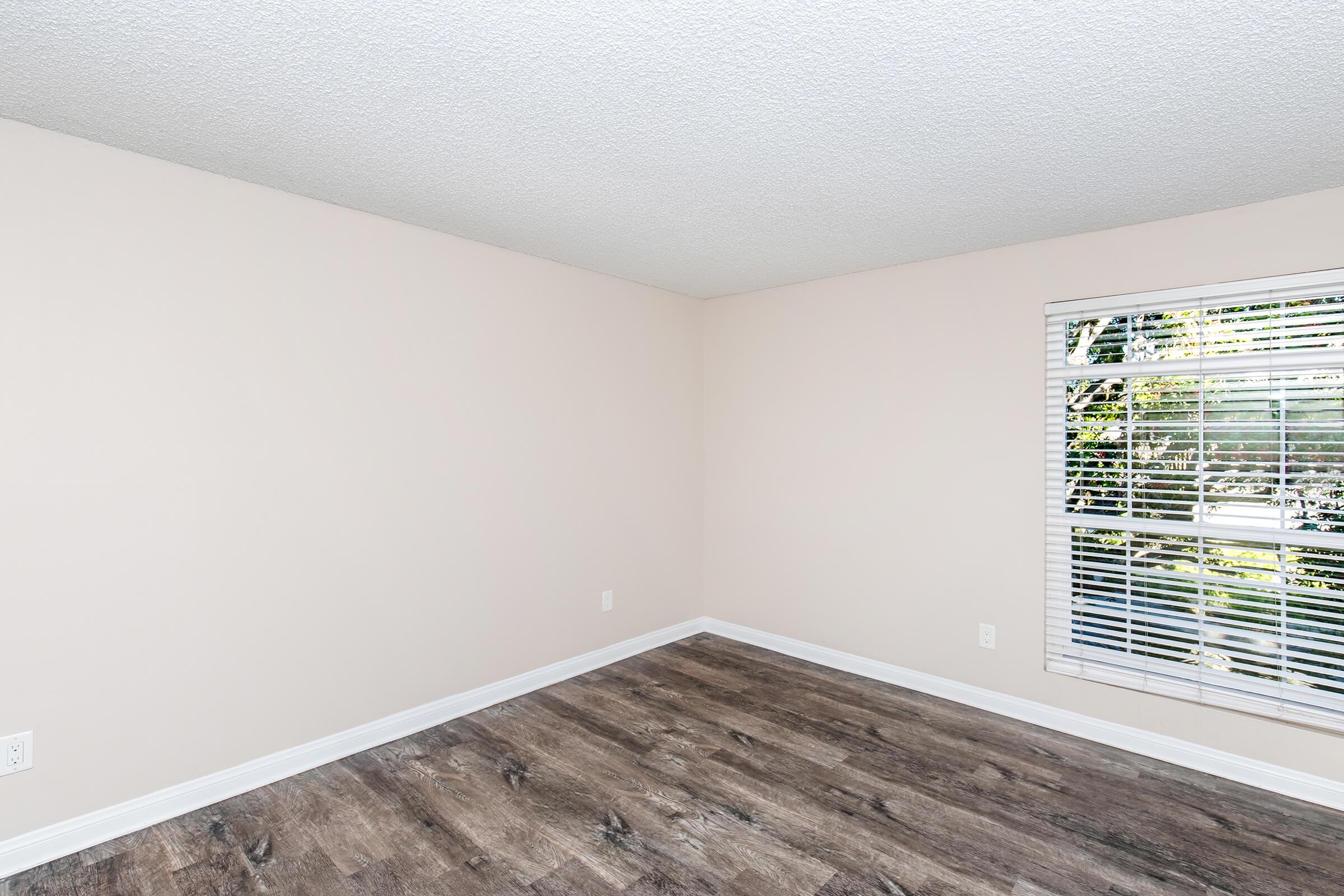 Empty room with light beige walls, white baseboards, and wooden flooring. A window with white blinds allows natural light to enter, casting shadows on the floor. The space is uncluttered and minimalistic, creating a neutral atmosphere suitable for various uses.