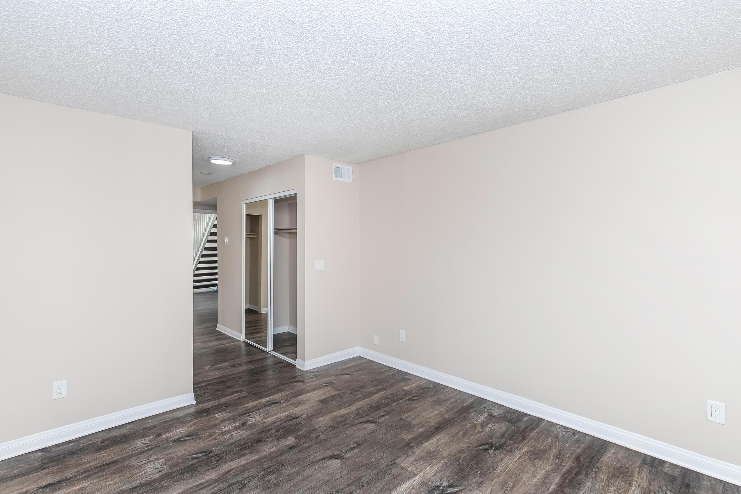 Interior view of a spacious, light-colored room featuring a textured ceiling, modern flooring, and a closet with sliding doors. The walls are painted in a soft beige, and there is a staircase visible in the background, adding to the open feel of the space.