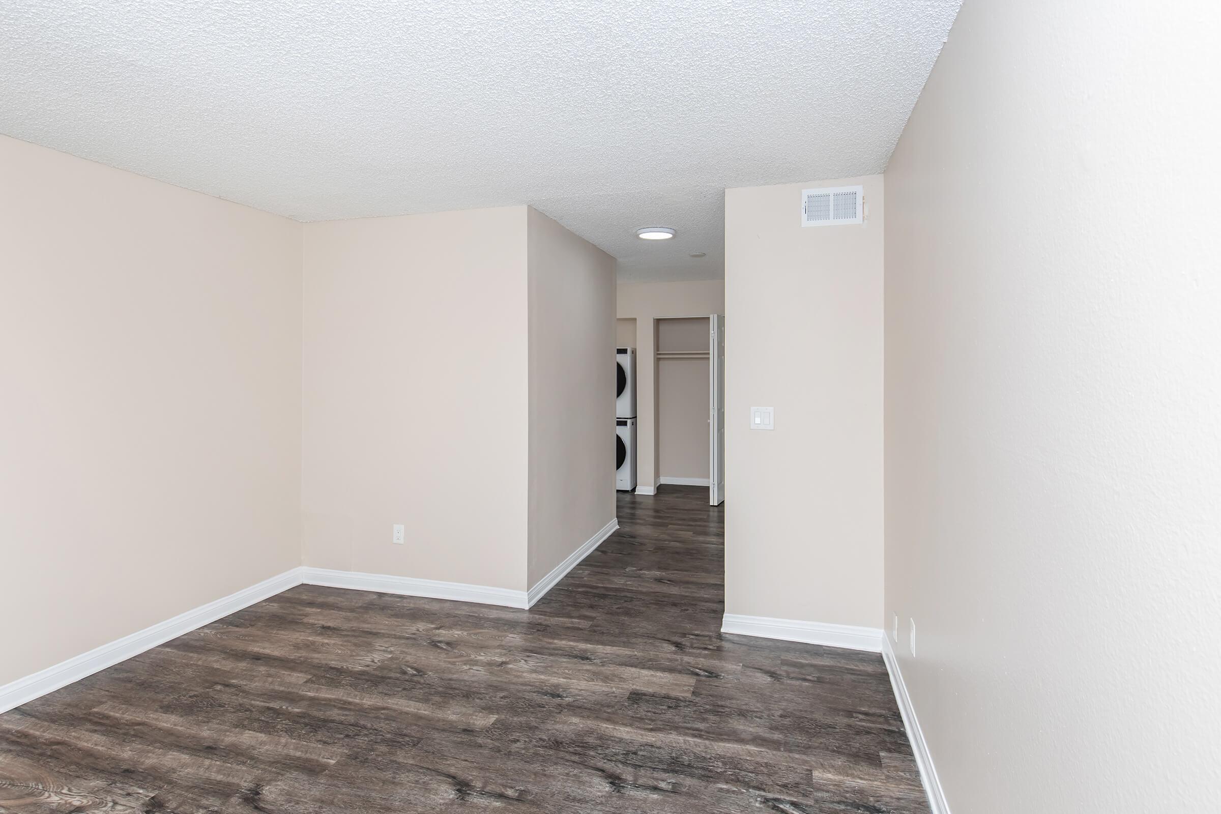 Empty interior of a room featuring light beige walls, a textured ceiling, and dark wood-like flooring. There is an open space leading to a closet area with a doorway on the left. The overall atmosphere is bright and spacious, with no furniture or decorations present.