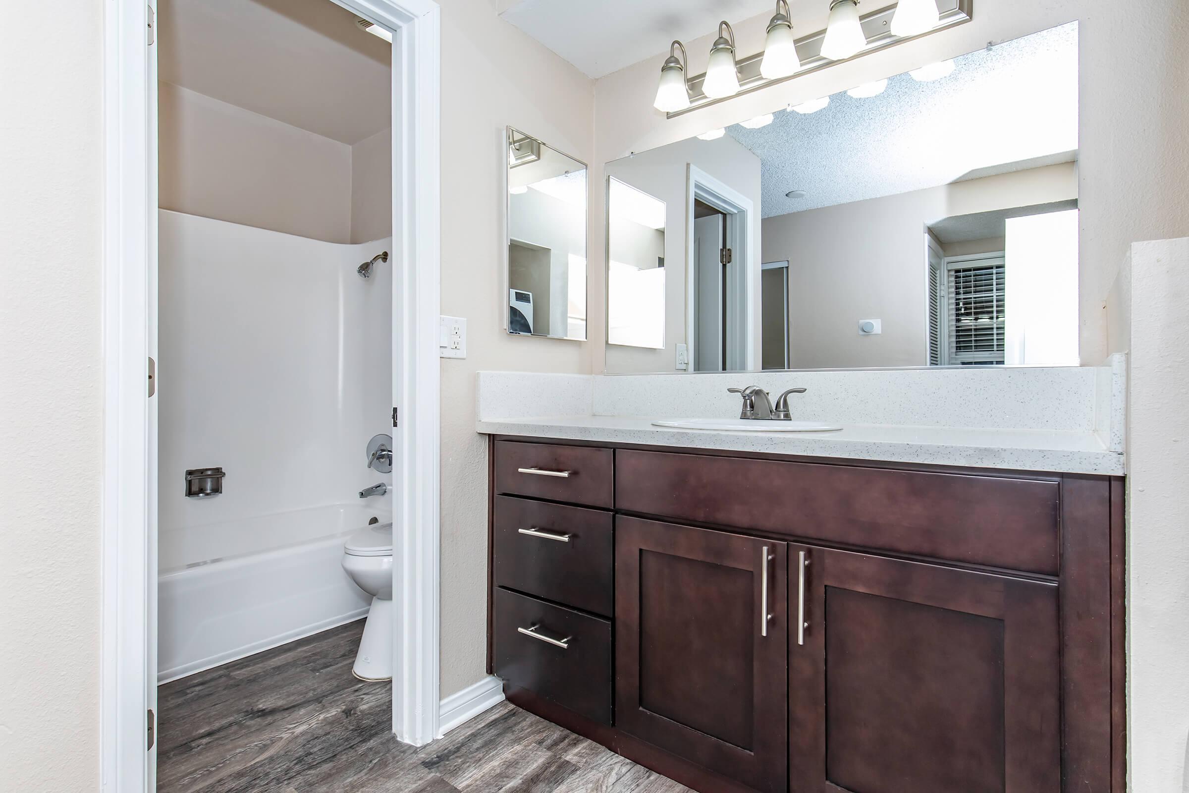 Modern bathroom featuring a white tub and shower combination, a contemporary vanity with dark wood cabinets, a sleek countertop, and a large mirror. The walls are painted in a light neutral shade, and the flooring is wood-like laminate. Soft lighting fixtures above the mirror provide a warm ambiance.