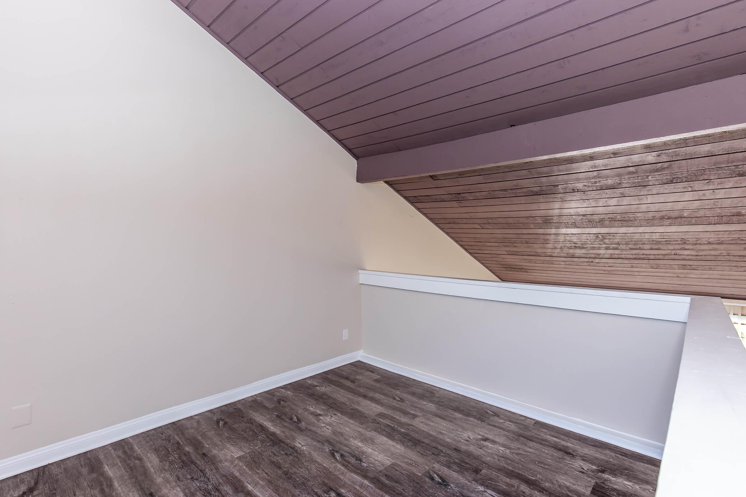 An empty room with a slanted ceiling, featuring beige walls and dark wooden flooring. The space has a white baseboard and a contrasting dark ceiling. The overall atmosphere is bright and minimalistic, creating a sense of openness.