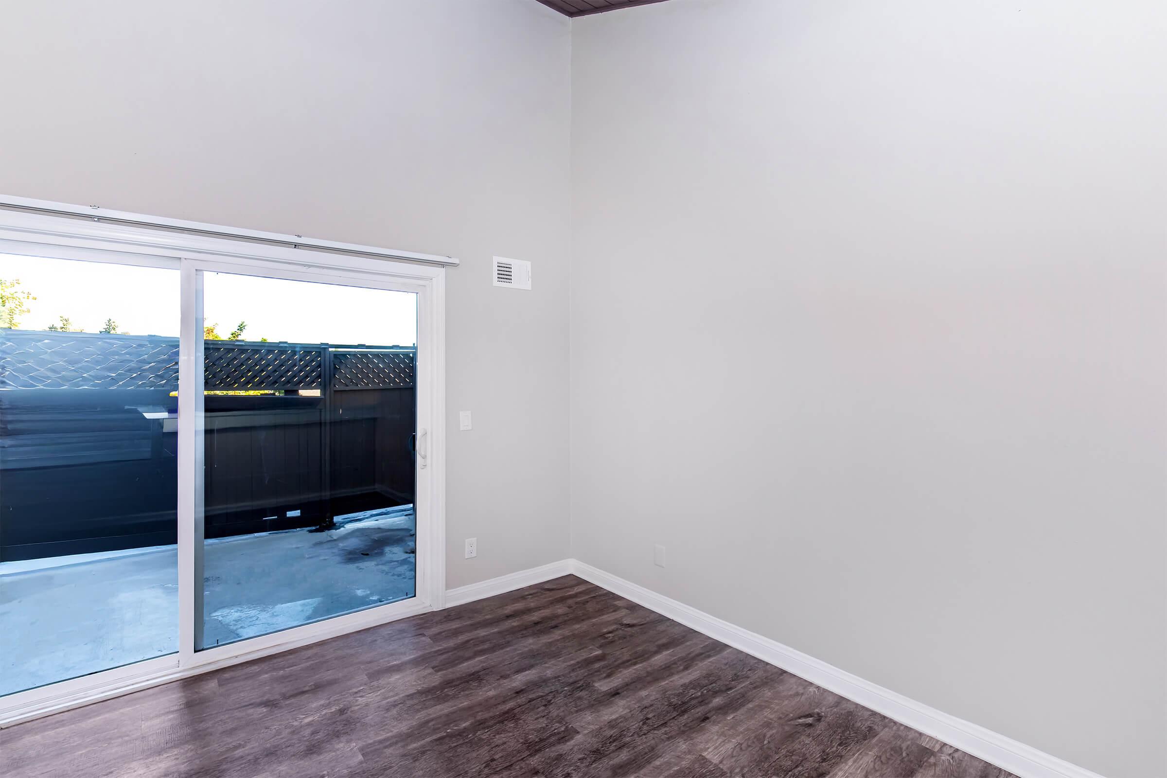 A vacant room with light-colored walls and a wooden floor. There is a large sliding glass door leading to an outdoor area with a balcony or patio. Natural light enters from outside, but the space is otherwise empty and lacking furniture or decor.