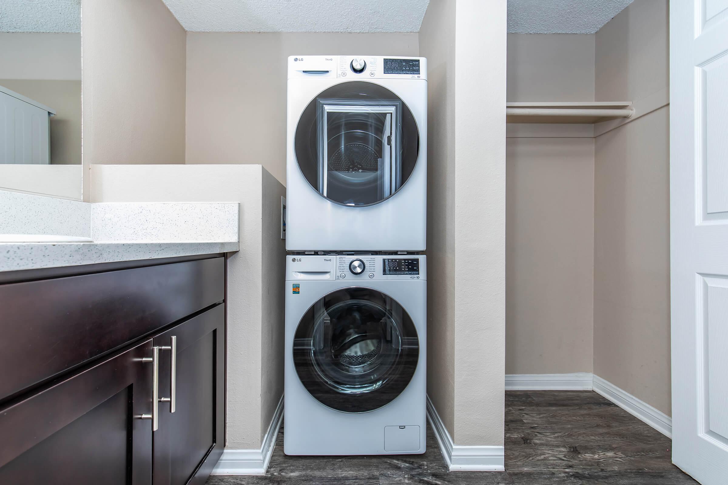 A modern laundry area featuring a stacked washer and dryer in sleek white. The appliances are situated against a light-colored wall, with a small countertop beside them. There is a closet space visible on the right, and the flooring is a dark wood laminate, providing contrast to the light walls.
