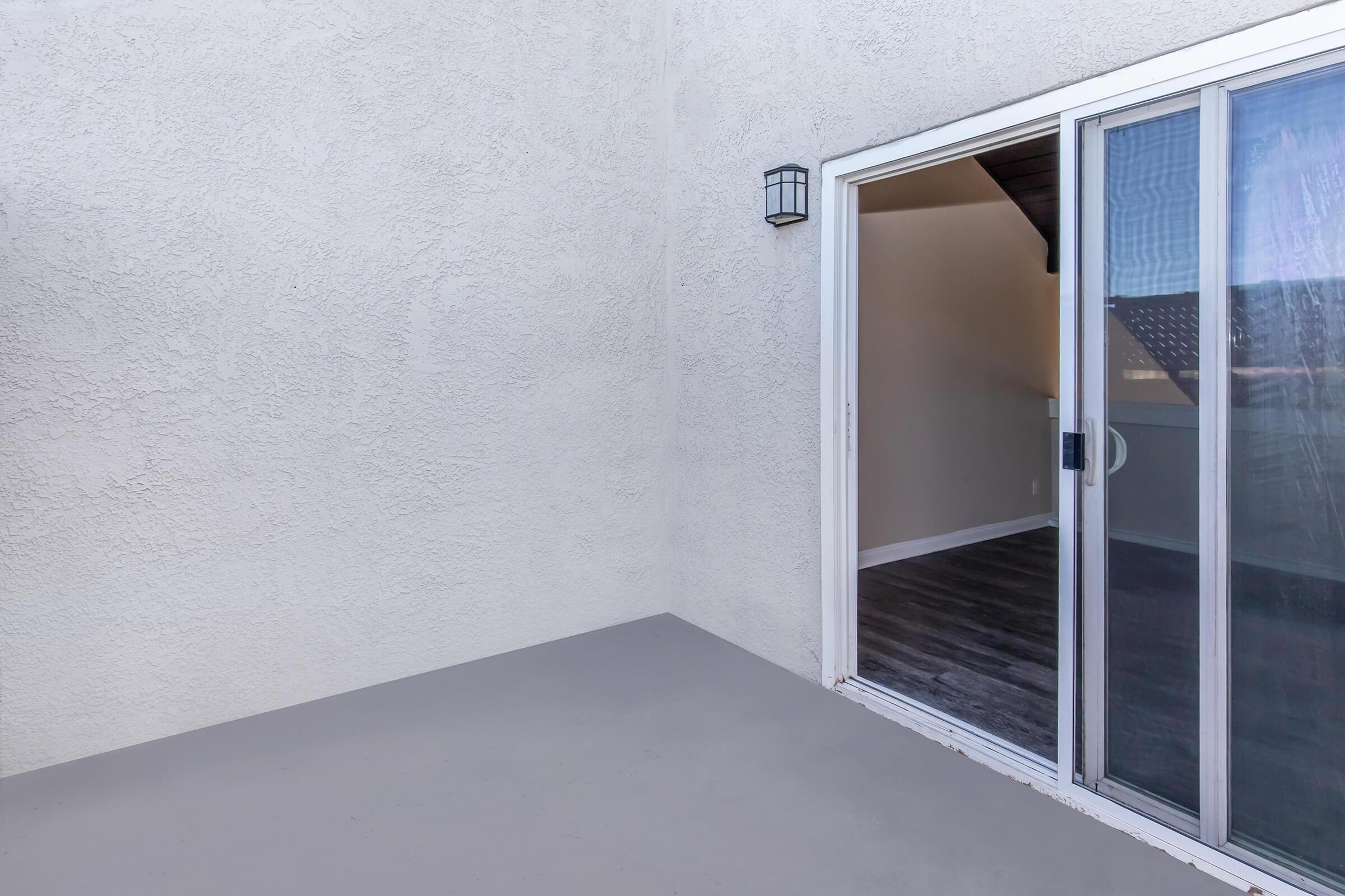 A partially open sliding glass door leads to a small patio area with a smooth gray surface. The wall is a light textured finish, and a wall-mounted light fixture is visible beside the door. The interior space beyond the door features light-colored walls and a wooden floor.