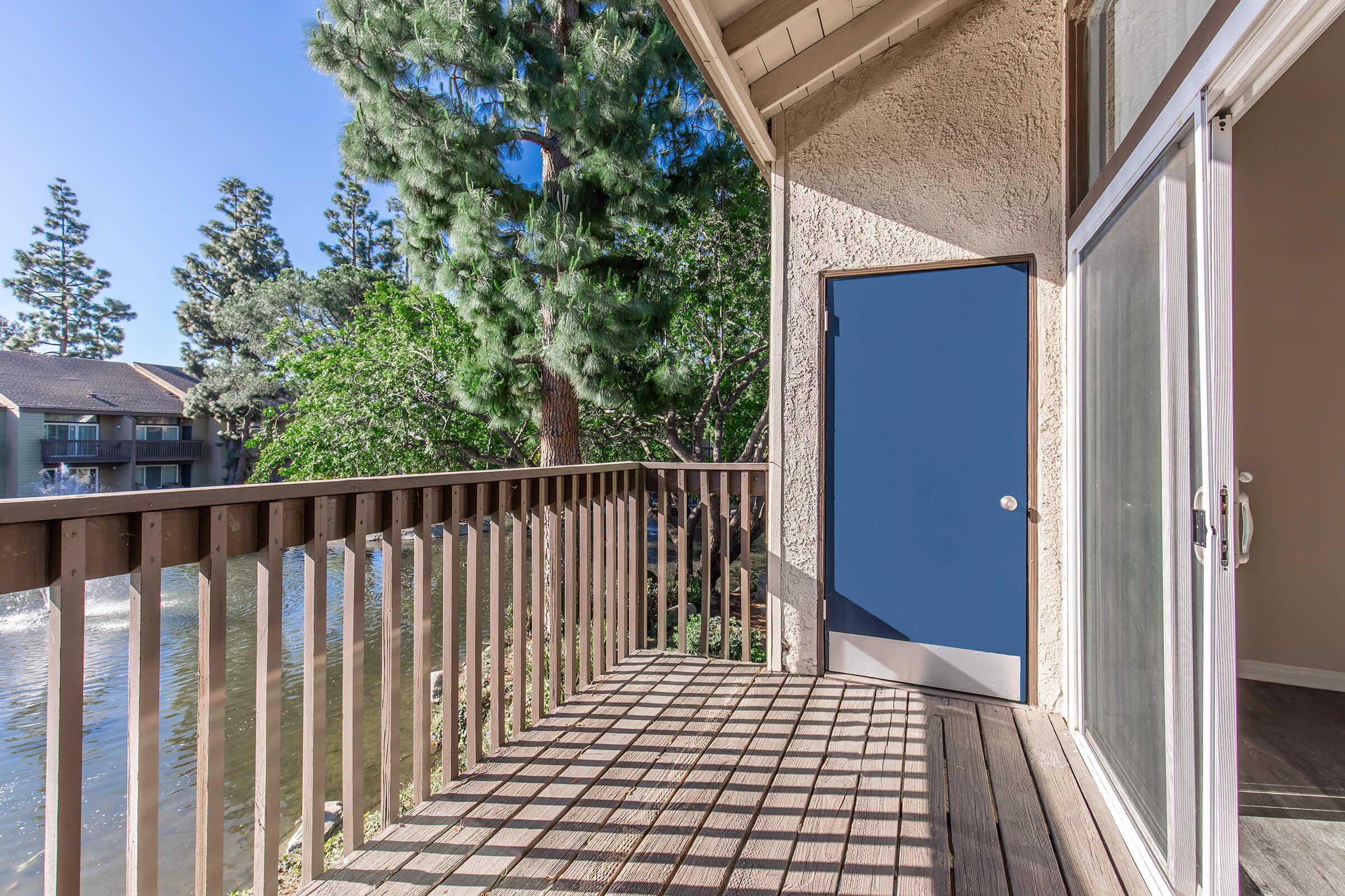A view from a balcony overlooking a calm body of water surrounded by trees. Sunlight casts shadows on the wooden deck, highlighting its texture. A closed blue door is visible, leading to the interior space. The scene conveys a peaceful, outdoor atmosphere in a residential area.