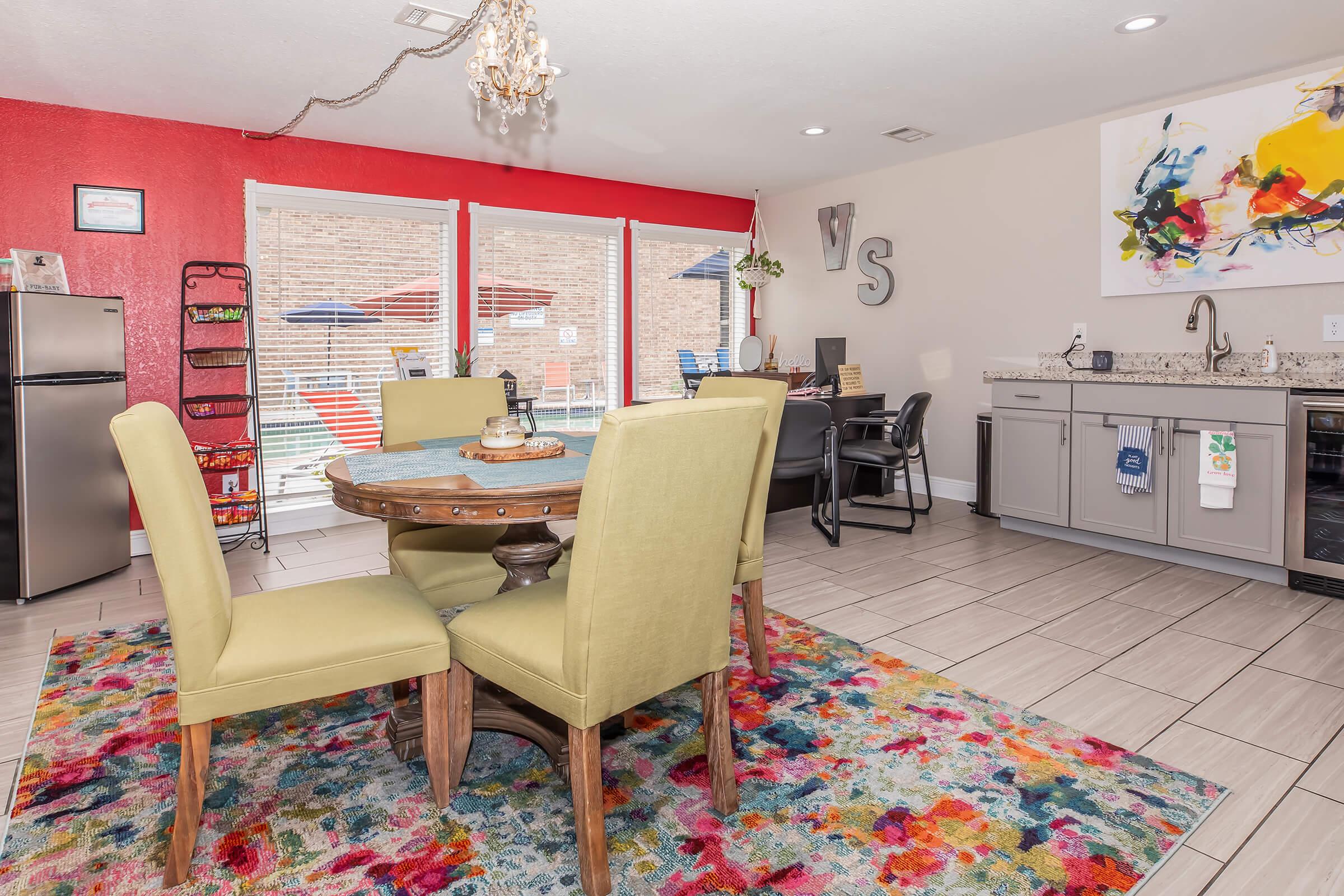 A bright and inviting kitchen-dining area featuring a round table with yellow chairs, a colorful rug, and modern appliances. The walls are painted in a mix of red and neutral tones, and large windows provide natural light. Decorative artwork and seating create a welcoming atmosphere.
