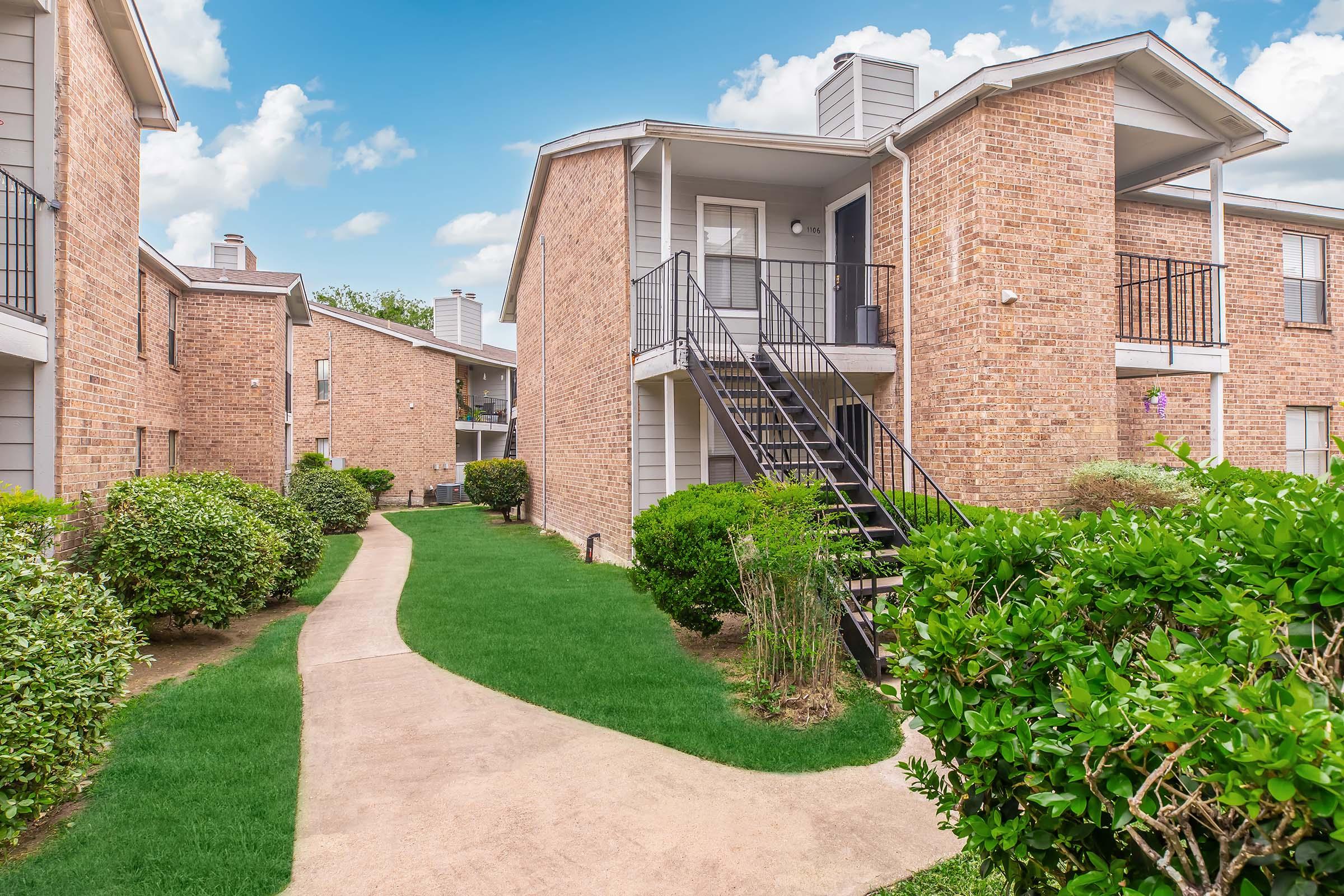 A well-maintained apartment complex featuring brick buildings with balconies, surrounded by neatly trimmed shrubs and green lawns. A concrete pathway leads through the landscape, connecting the buildings under a bright blue sky with fluffy white clouds.