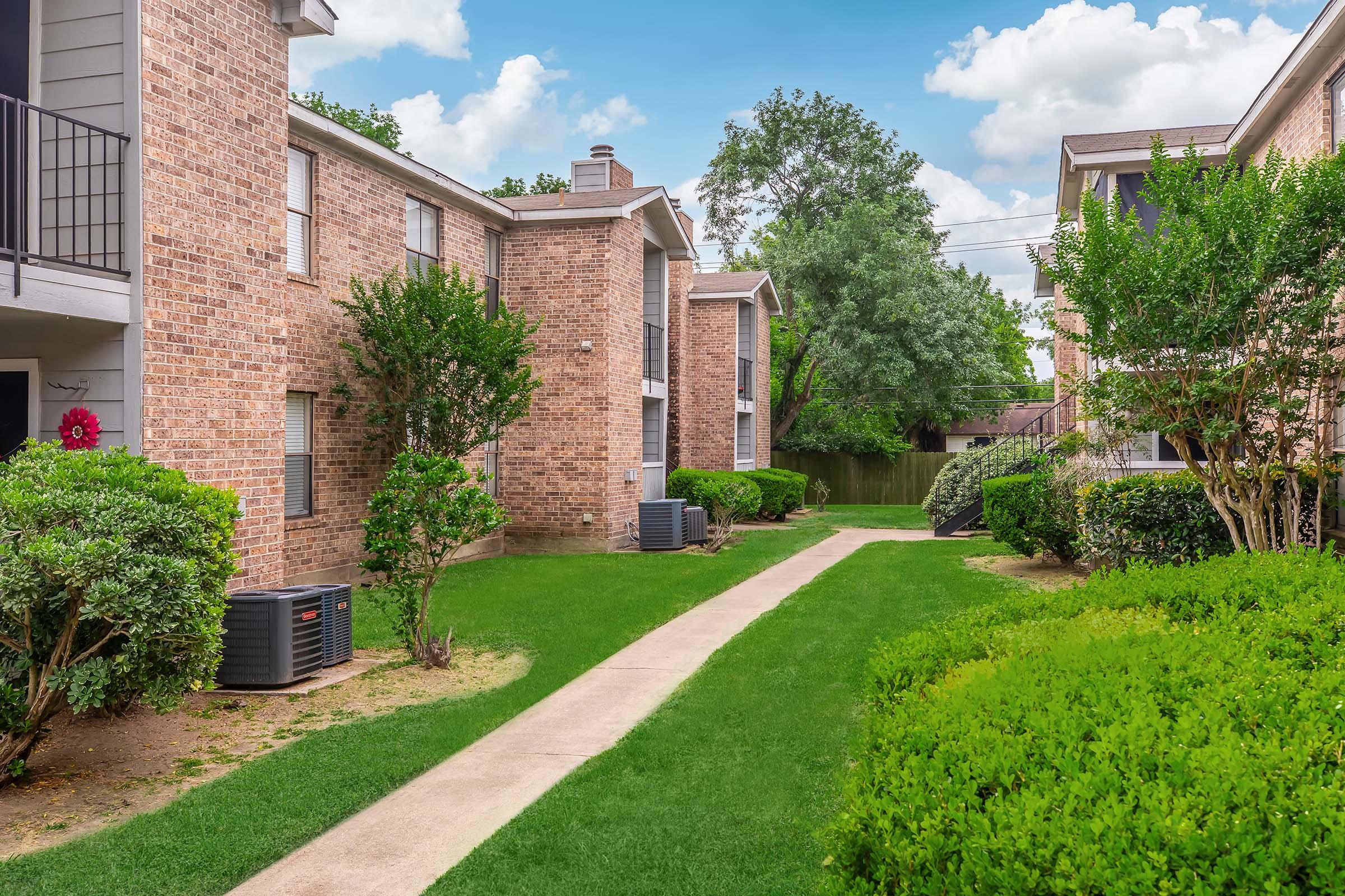 A view of a landscaped courtyard in an apartment complex, featuring brick buildings, green grass, and well-maintained shrubs. Pathways wind through the greenery, with air conditioning units visible at the base of the buildings under a partly cloudy sky.
