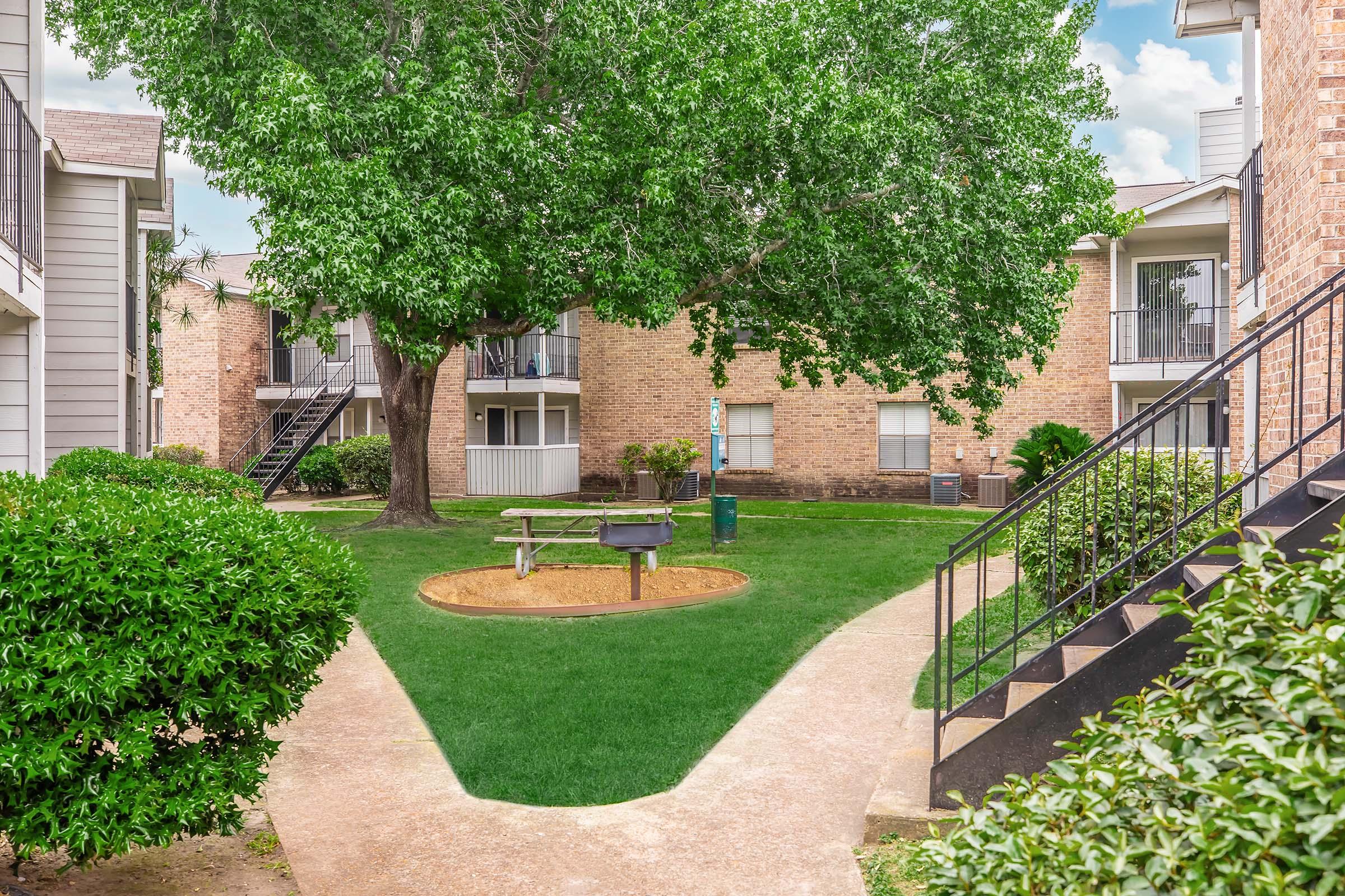 A grassy courtyard surrounded by apartment buildings, featuring a large tree, picnic table, and a barbecue grill. Pathways lead through manicured bushes towards stairs of the buildings. The sky is partly cloudy, creating a bright and inviting atmosphere.