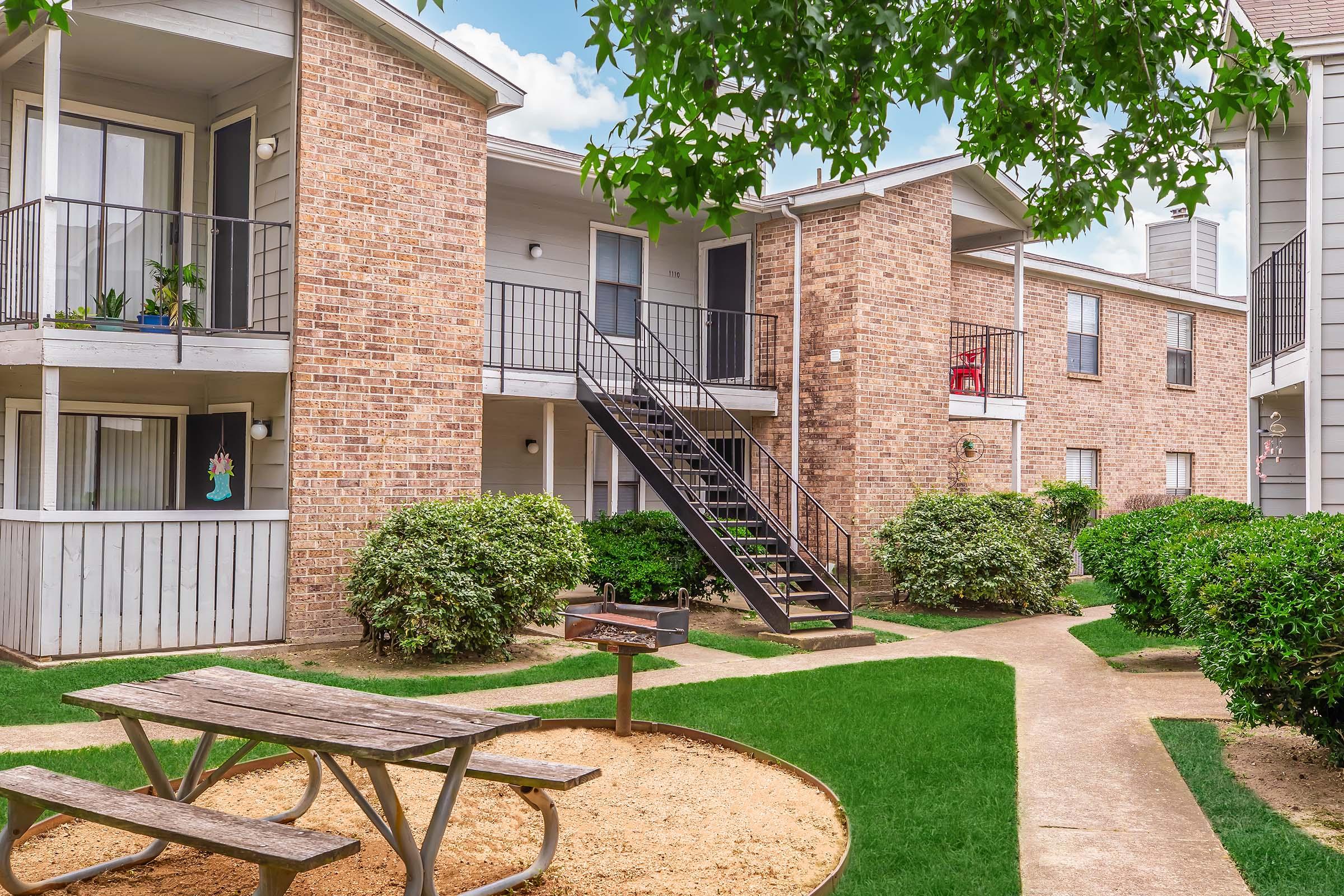 A view of a residential apartment complex featuring brick buildings. There are balconies, a staircase, and well-maintained greenery. A picnic table is set on a sandy area, with a path winding through the landscaped grounds. The sky is bright, adding to the inviting atmosphere.