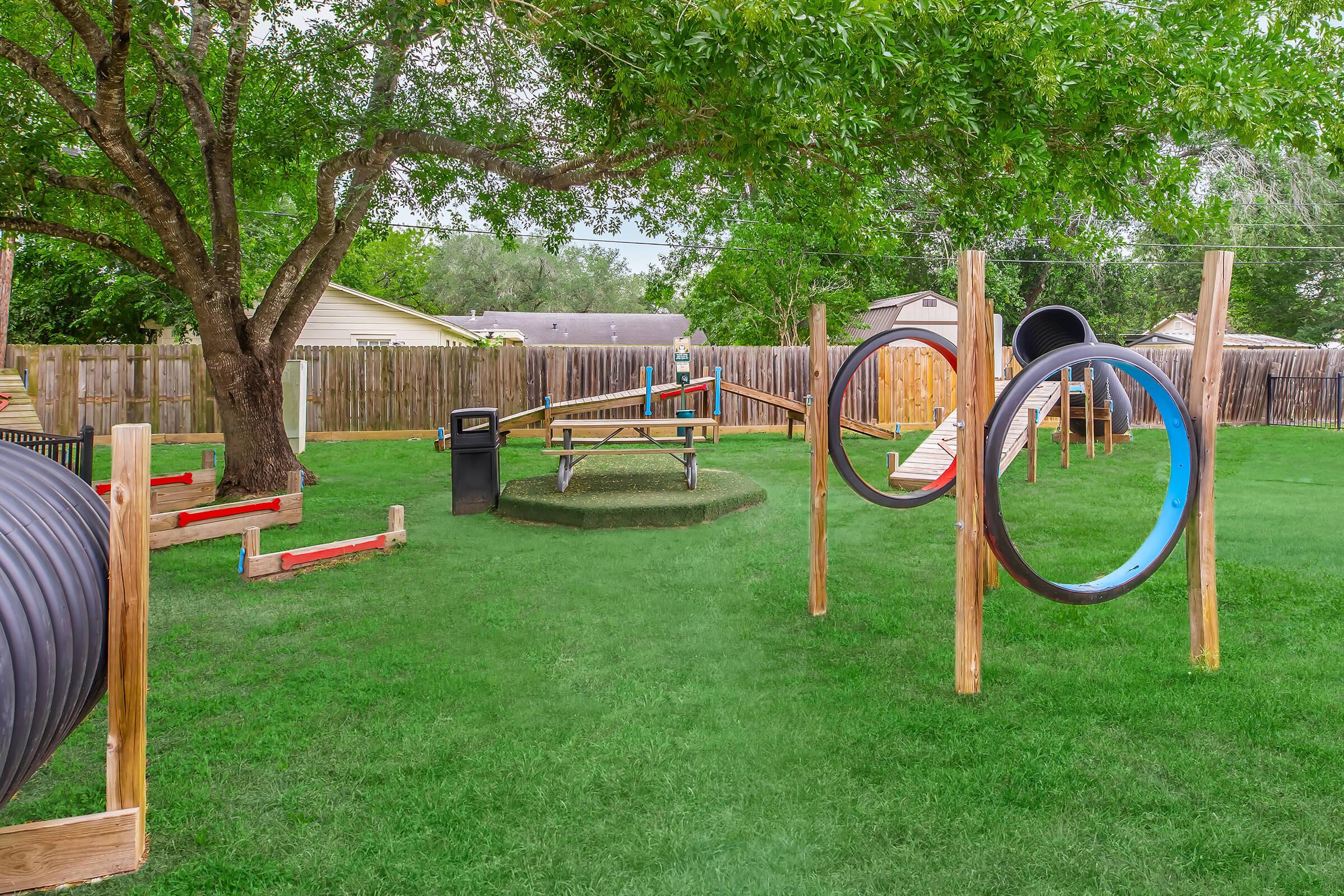 A playground scene featuring various structures, including a slide, climbing equipment, and circular tunnels. The area is grassy with trees surrounding the playground, and there's a waste bin nearby. A wooden platform serves as a central feature, creating a vibrant, inviting outdoor space for children to play.