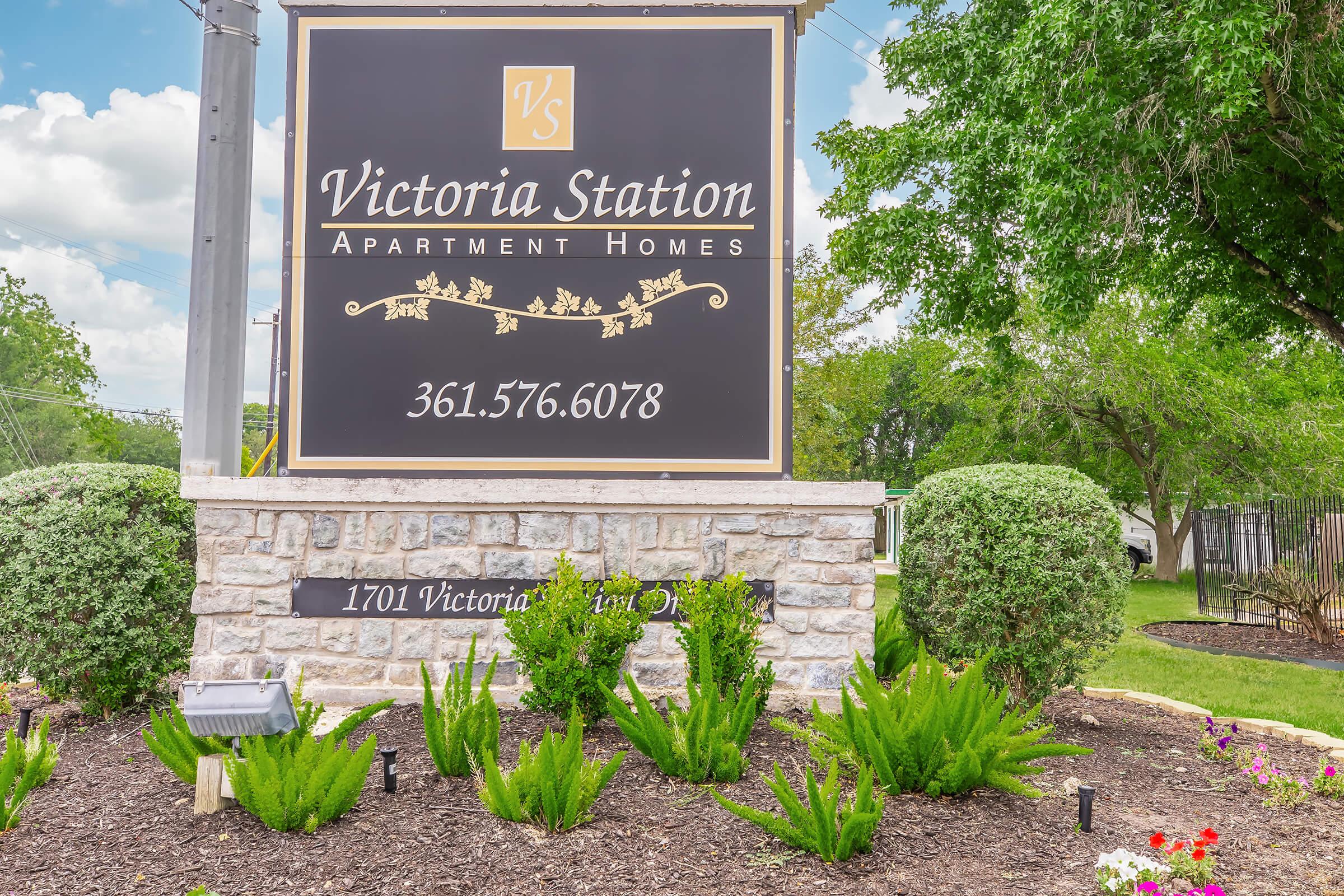 Sign for Victoria Station Apartment Homes, featuring the text "Victoria Station" and the phone number 361.576.6078. The sign is framed by greenery and decorative landscaping, with a stone base. The location address is displayed as "1701 Victoria St, Unit D." Bright blue sky with fluffy clouds in the background.