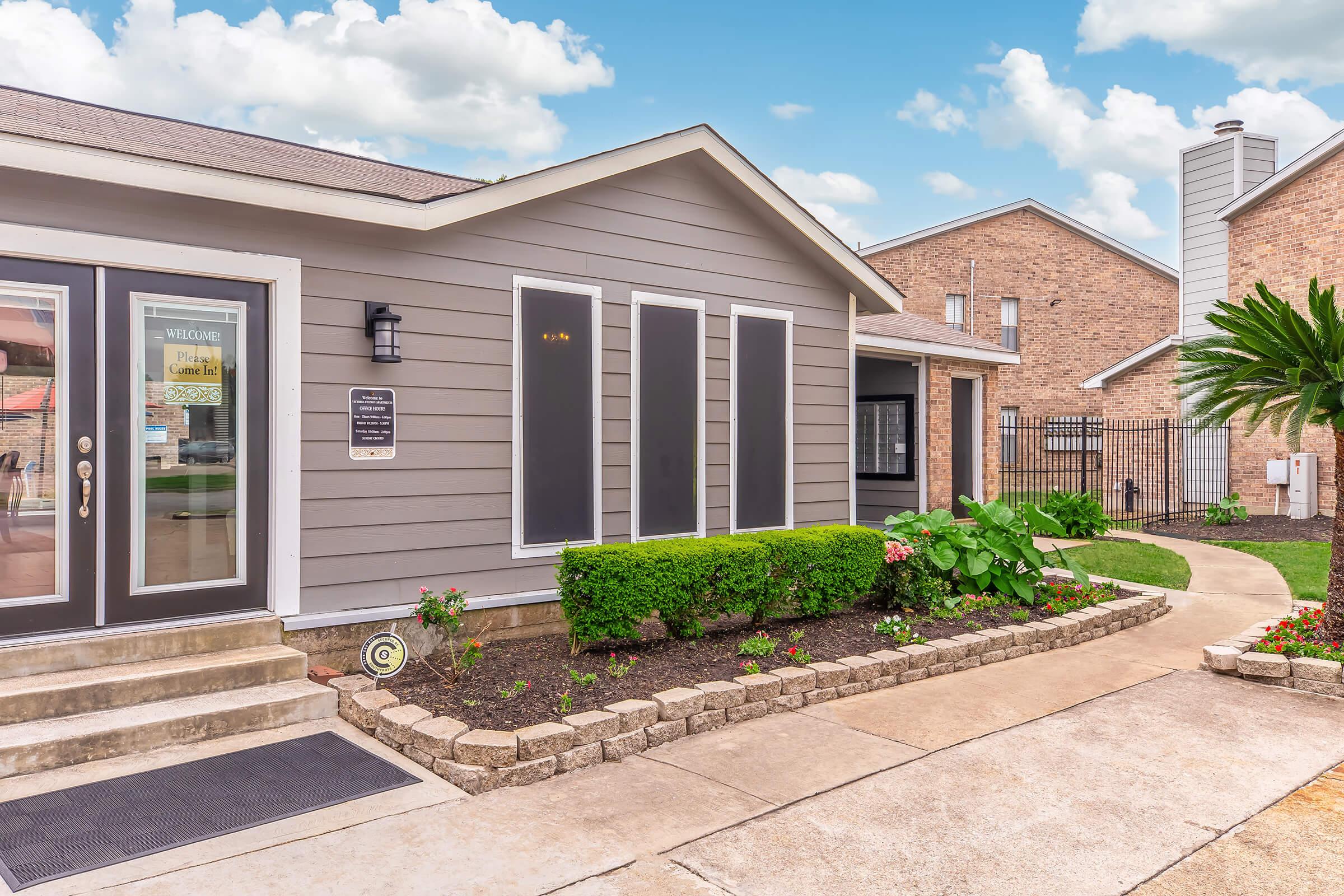 A modern gray house with multiple large windows and a welcoming front entrance. There is a well-maintained garden with green plants and shrubs, curving stone pathways, and a brick building visible in the background. The sky is bright with fluffy clouds, creating a pleasant atmosphere.