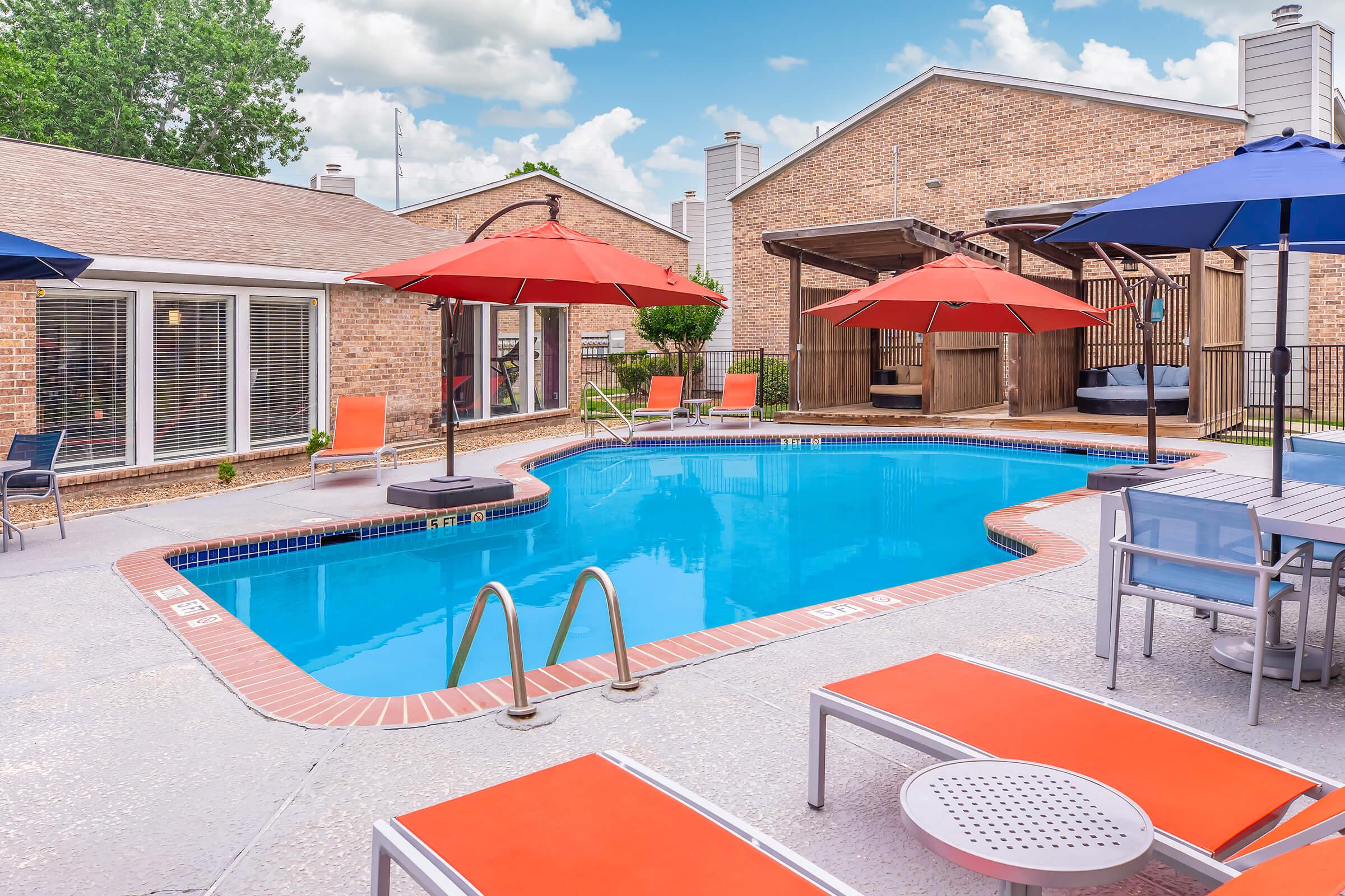 A bright blue swimming pool surrounded by red parasols and lounge chairs. Nearby, there are shaded seating areas with wooden structures. The backdrop features brick buildings and a clear sky with fluffy white clouds, creating a vibrant, inviting atmosphere.