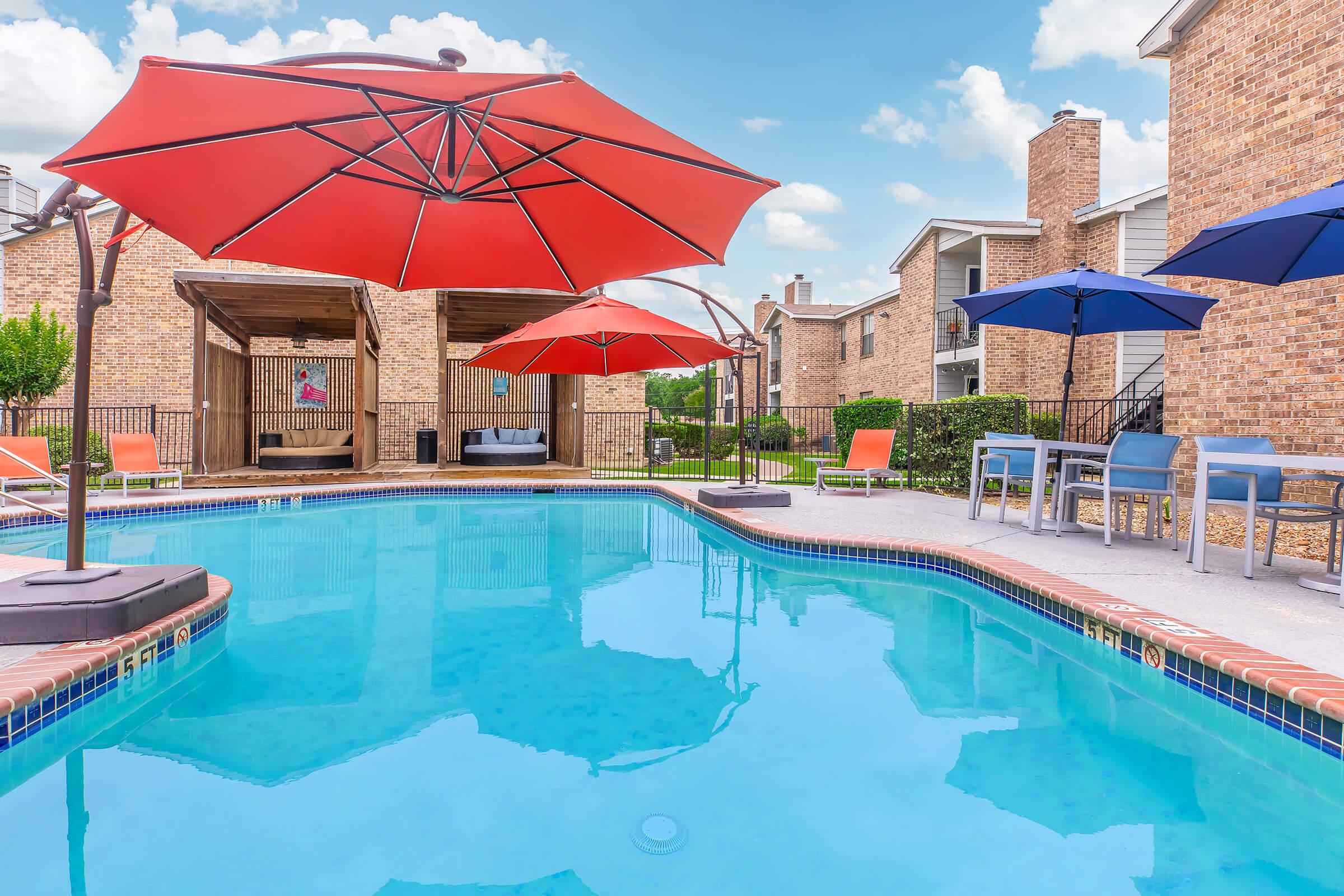 A swimming pool surrounded by lounge chairs and large red and blue umbrellas. In the background, there are two wooden pavilions and brick apartment buildings. The sky is bright with fluffy clouds, creating a relaxing outdoor atmosphere.