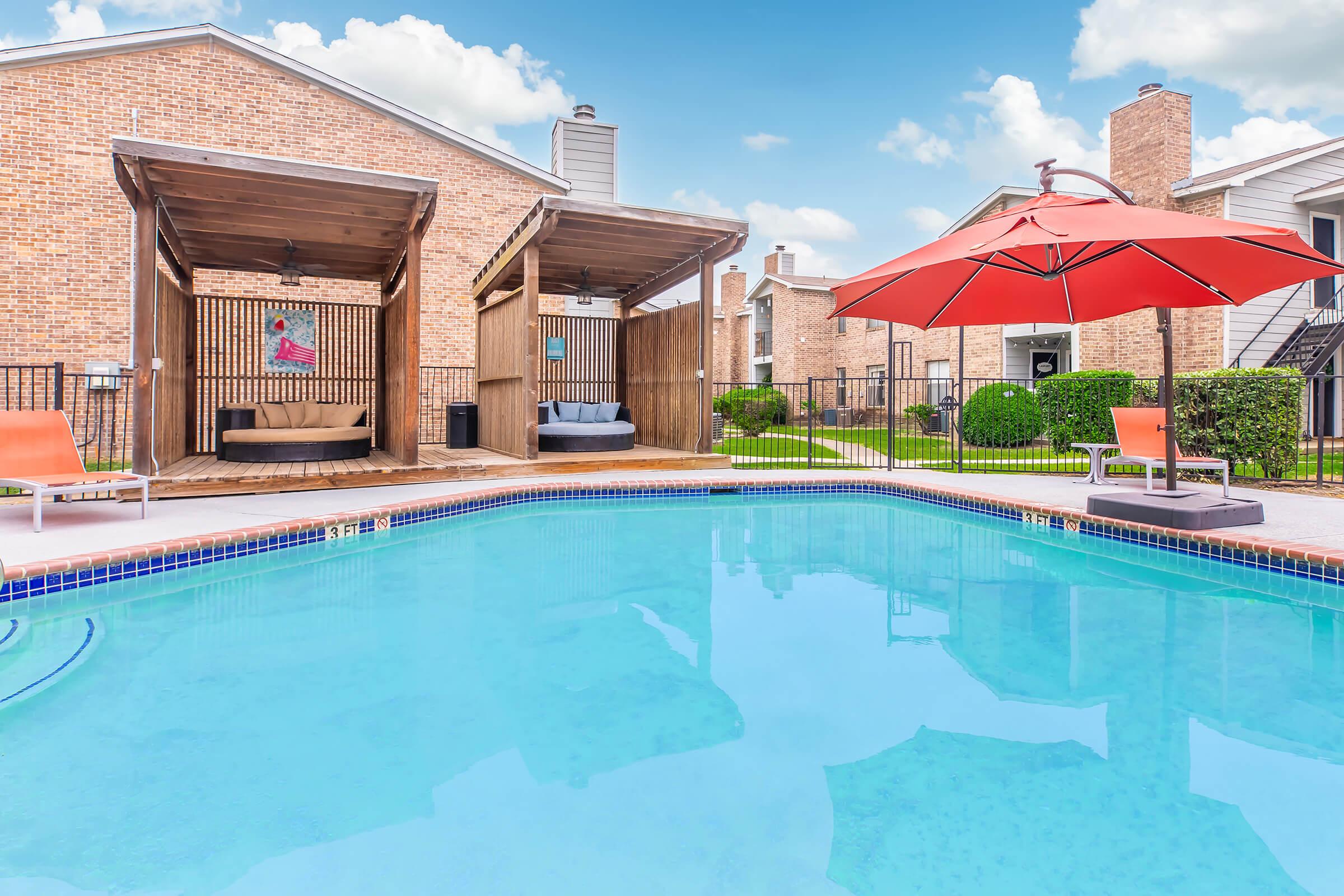 A clear swimming pool with a wooden deck, surrounded by two cabanas and an umbrella. In the background, there are brick buildings and well-maintained greenery. The sky is blue with some clouds, creating a pleasant atmosphere. Perfect for relaxation and leisure by the poolside.