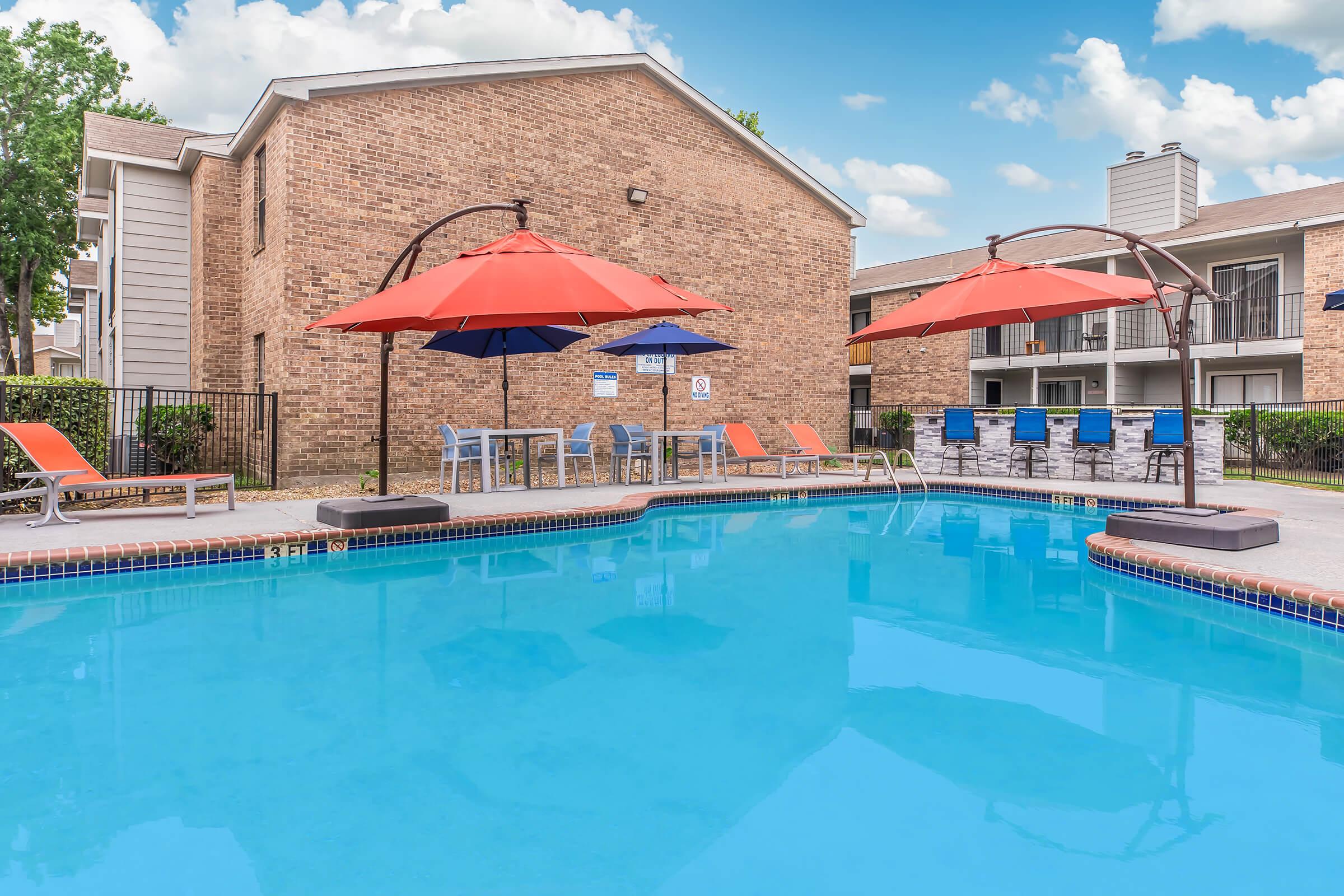 A swimming pool surrounded by lounge chairs and colorful umbrellas. The pool water is bright blue, and there are several blue chairs arranged nearby. The backdrop features a brick apartment building with multiple floors and green trees under a clear blue sky.