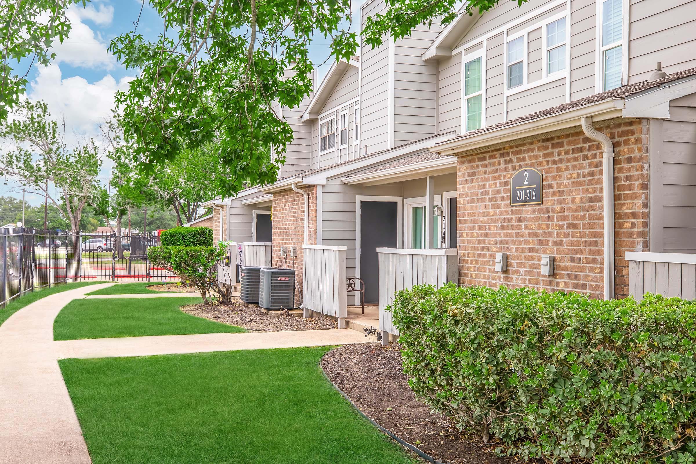 A well-maintained apartment complex featuring beige and gray siding. The pathway leads through a lush green lawn, bordered by manicured shrubs. Each unit has a small porch area, with a few air conditioning units visible. Trees and blue skies create a pleasant atmosphere.