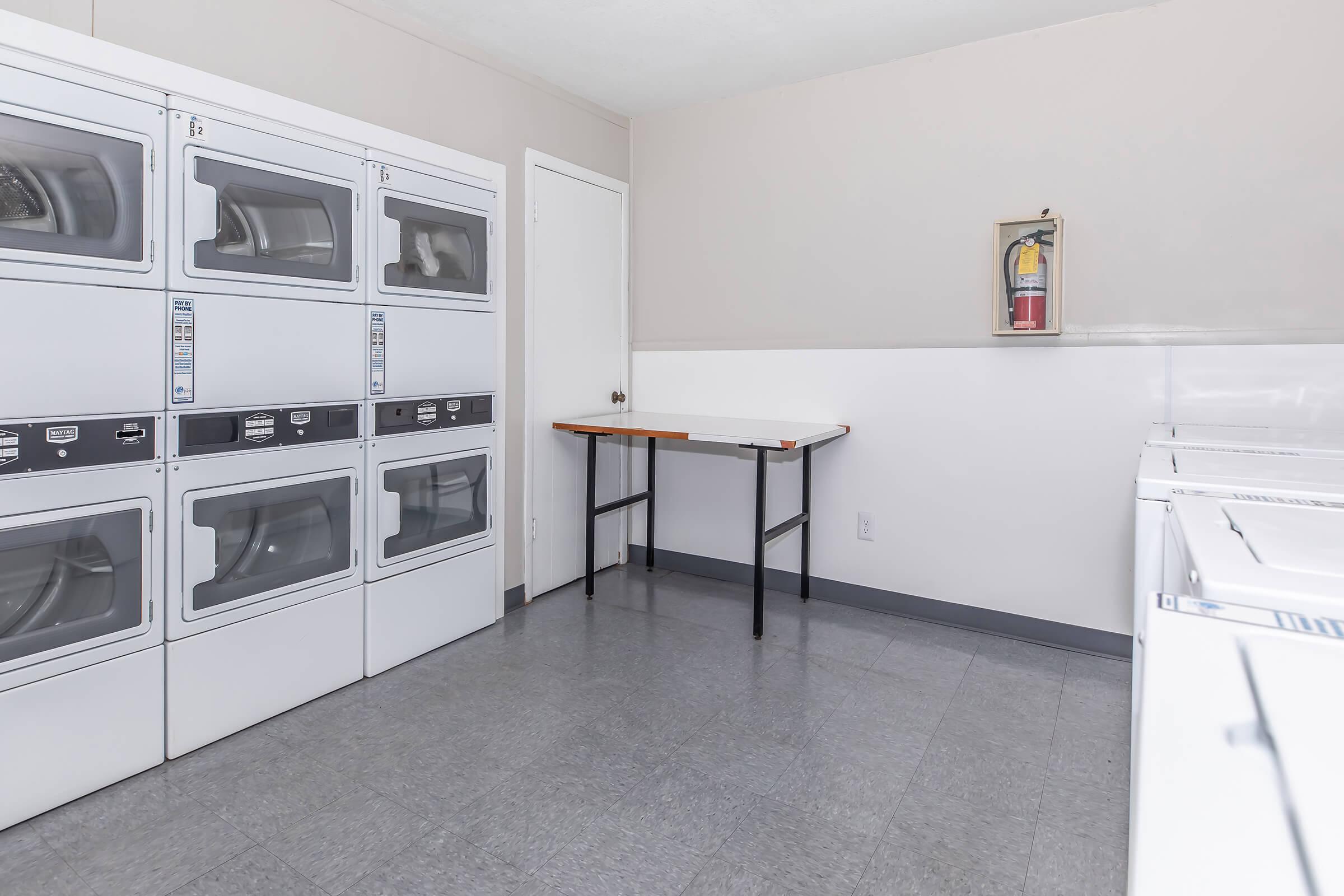A clean, well-lit laundry room featuring several stacked washing machines and dryers on the left. A simple wooden table is positioned in the center, and a fire extinguisher is mounted on the wall in the corner. The walls are light-colored, and the floor is a gray tile.