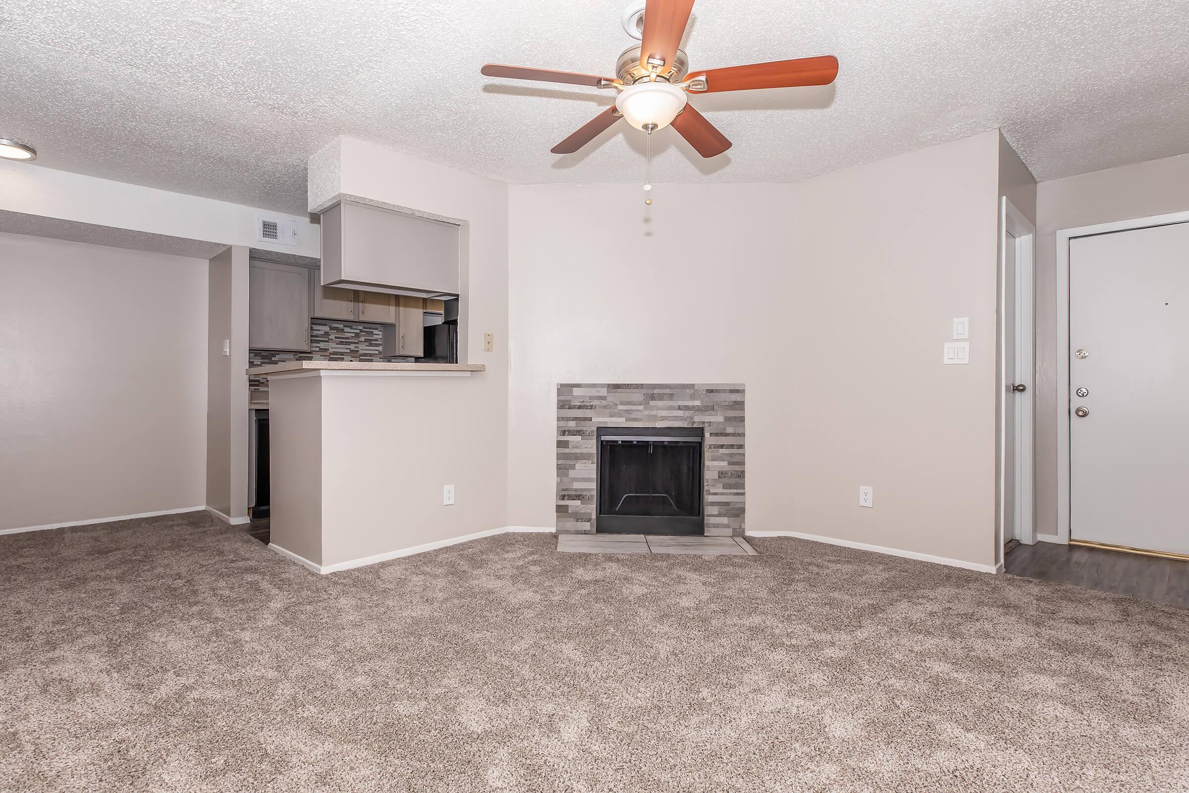 Living room featuring a ceiling fan, carpeted flooring, and a decorative fireplace. There's an open layout leading to a modern kitchen with a bar counter. Neutral-colored walls enhance the spacious feel, and a doorway visible on the right leads outside.