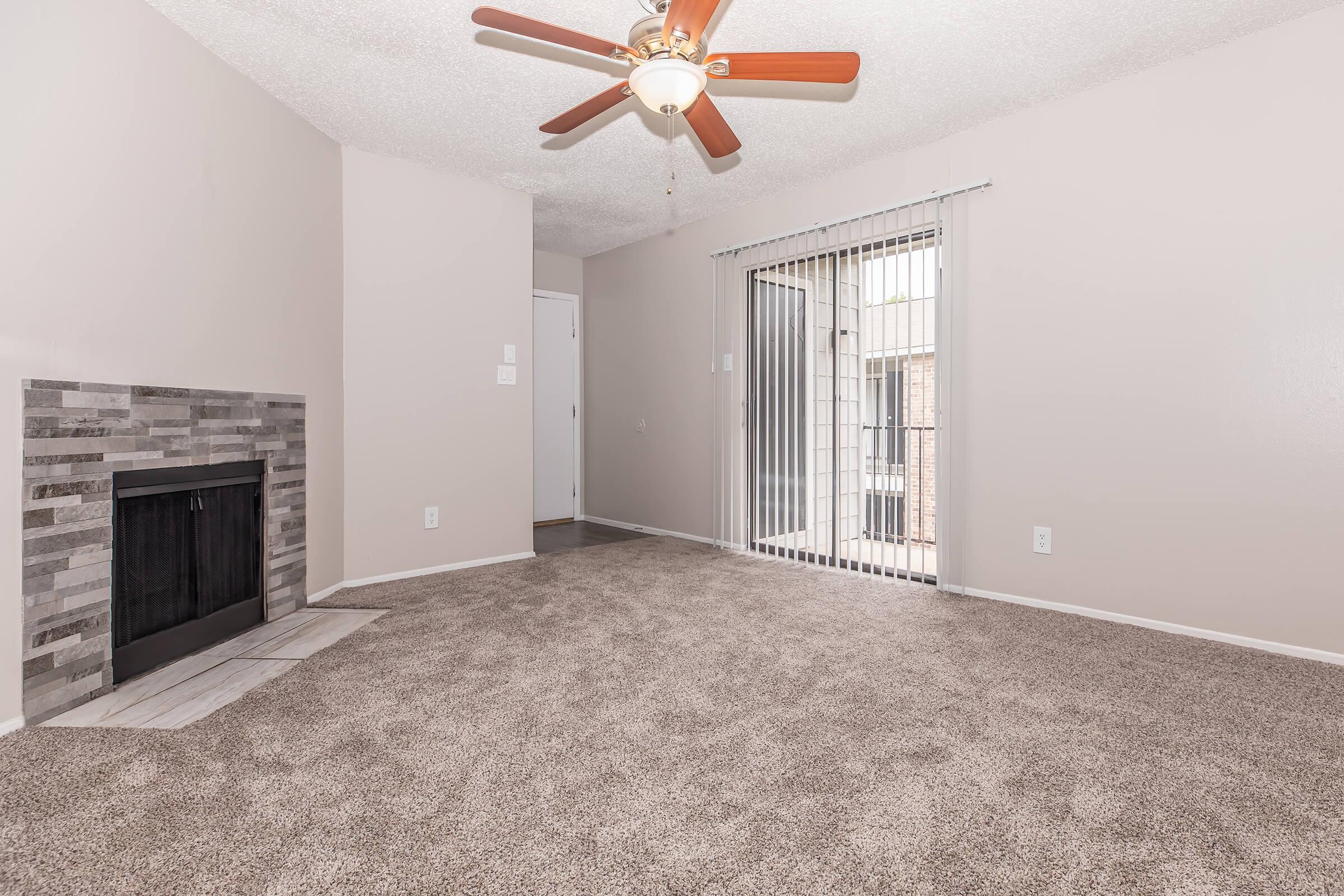 A spacious living room featuring a ceiling fan, carpeted flooring, and a decorative fireplace with stone accents. Large sliding glass doors lead to a balcony, allowing natural light to fill the room. The walls are painted in a light color, and the layout is open and inviting.
