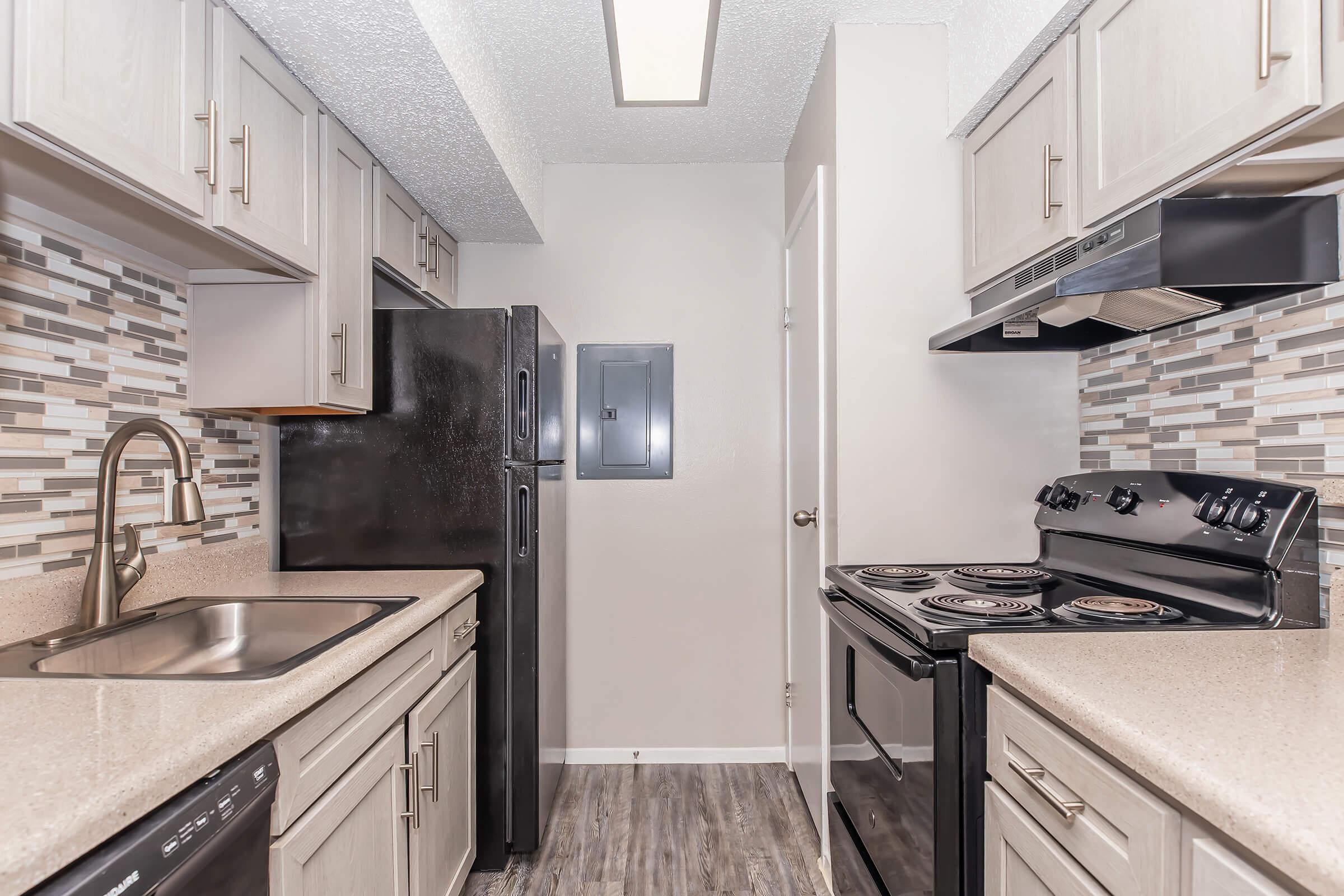 A modern kitchen featuring a black refrigerator and stove, light-colored cabinets, and a countertop with a textured finish. The backsplash consists of decorative tiles in neutral tones. The kitchen also includes a stainless steel sink and ample cabinet storage.