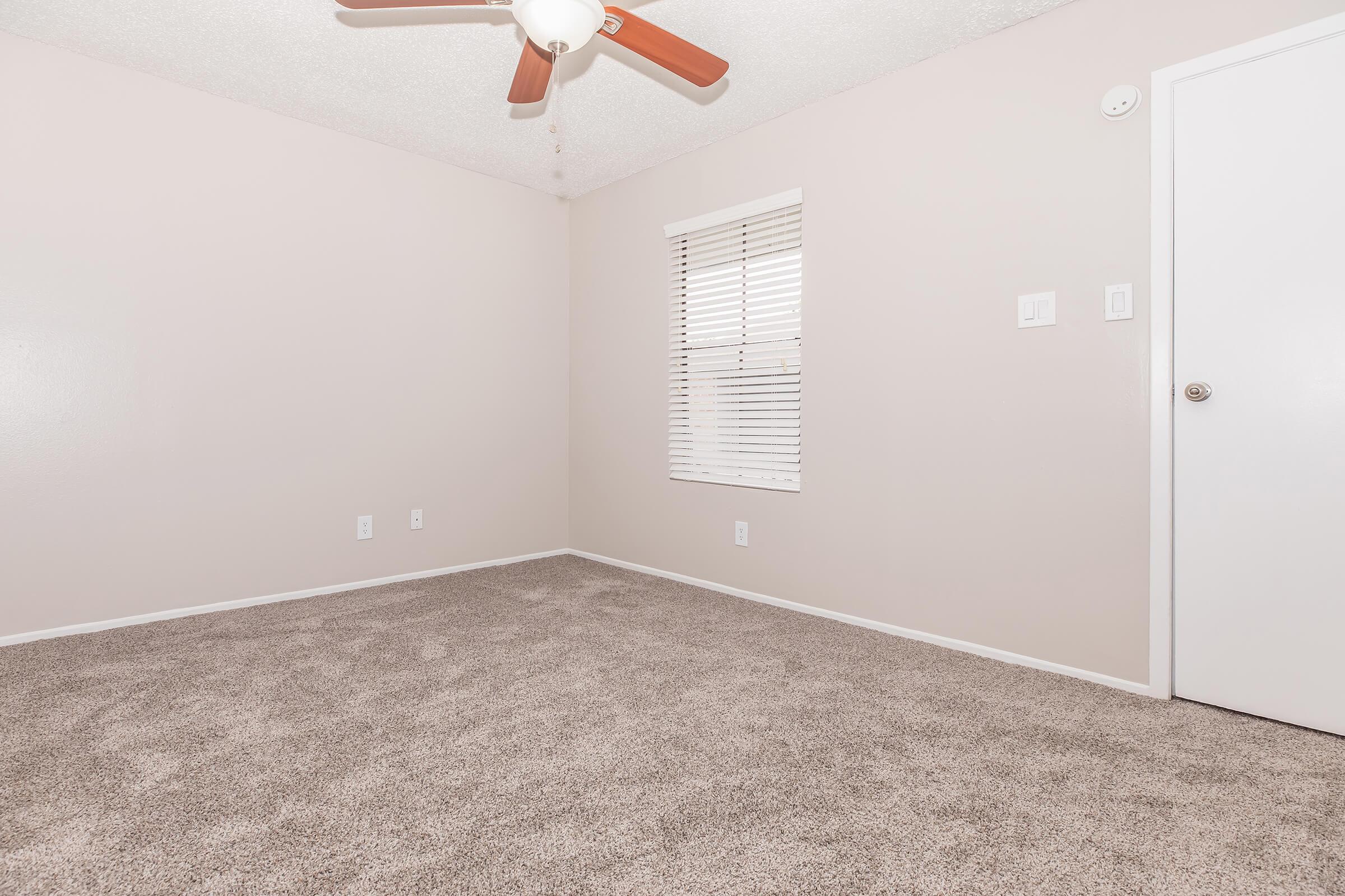 Empty room with light beige walls and carpet. Features a ceiling fan with wooden blades, a window with horizontal blinds, and a white door. The room is well-lit, but minimal furnishings give it a spacious feel.