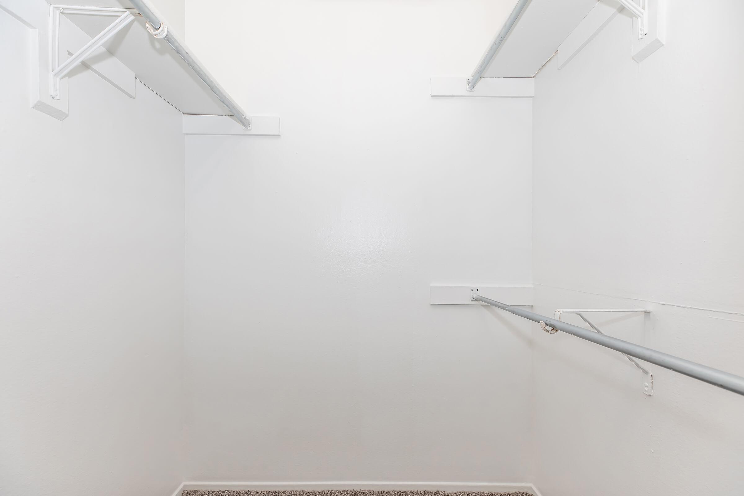 Empty closet with white walls and shelves, featuring two hanging rods on opposite sides. The floor is carpeted, and there are no visible items or clothing in the space.