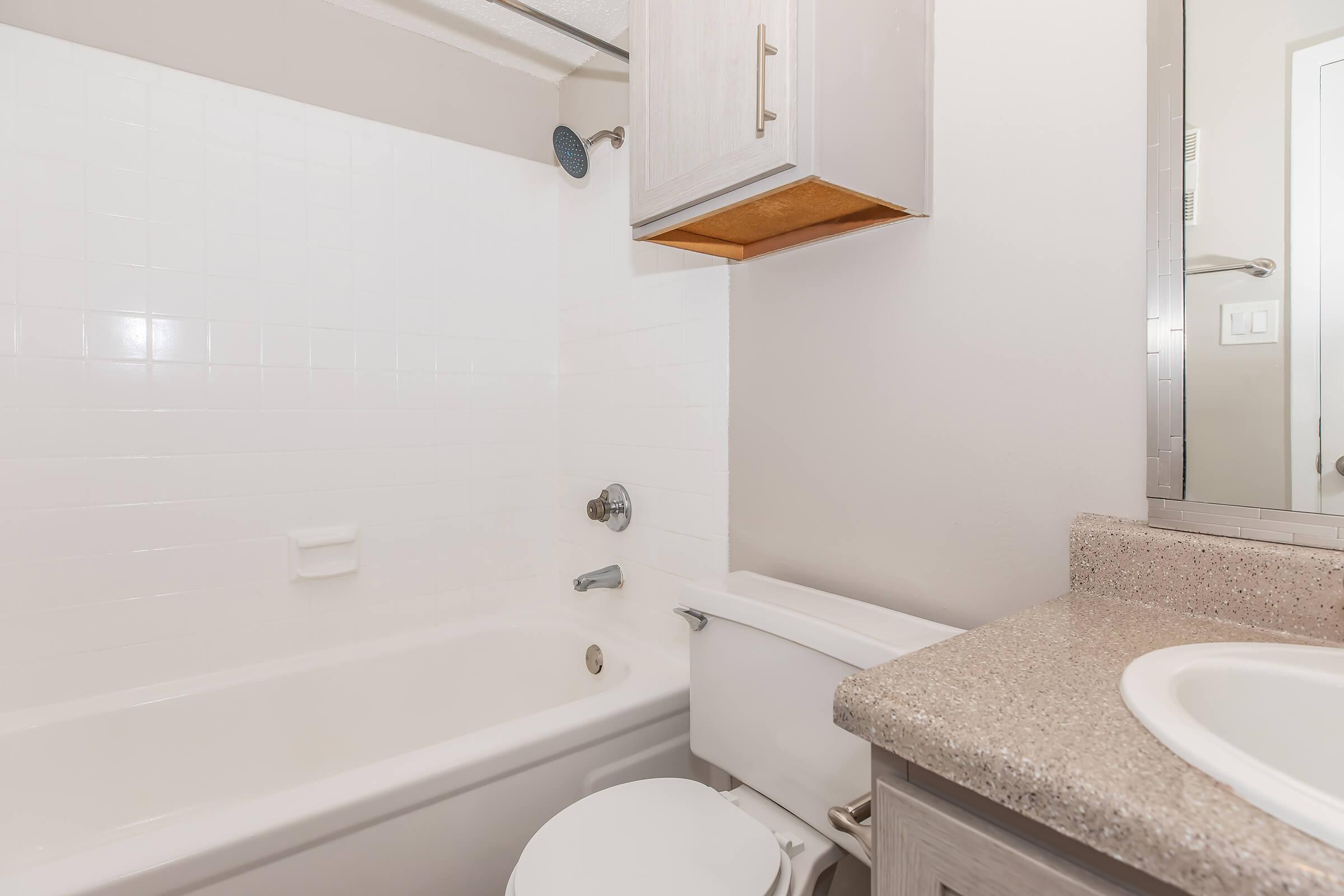 A clean bathroom featuring a white bathtub, a showerhead, a toilet, and a sink with a granite countertop. The walls are tiled and painted in neutral tones, and there is a mirror above the sink. A wooden cabinet is mounted above the toilet for storage.