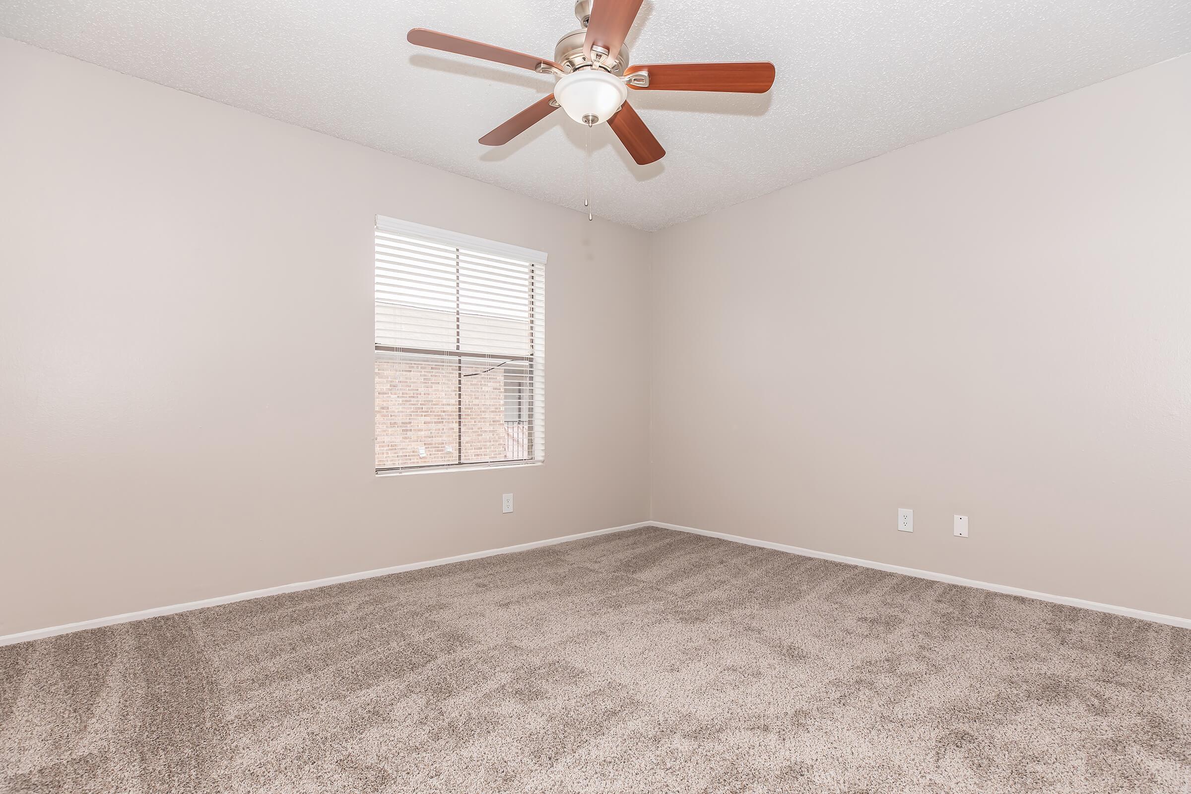 A spacious, empty room with light beige walls and a ceiling fan with wooden blades. The floor is covered in plush, light-colored carpet. A window with blinds allows natural light to enter, illuminating the space. The room is uncluttered, creating a serene atmosphere suitable for various purposes.