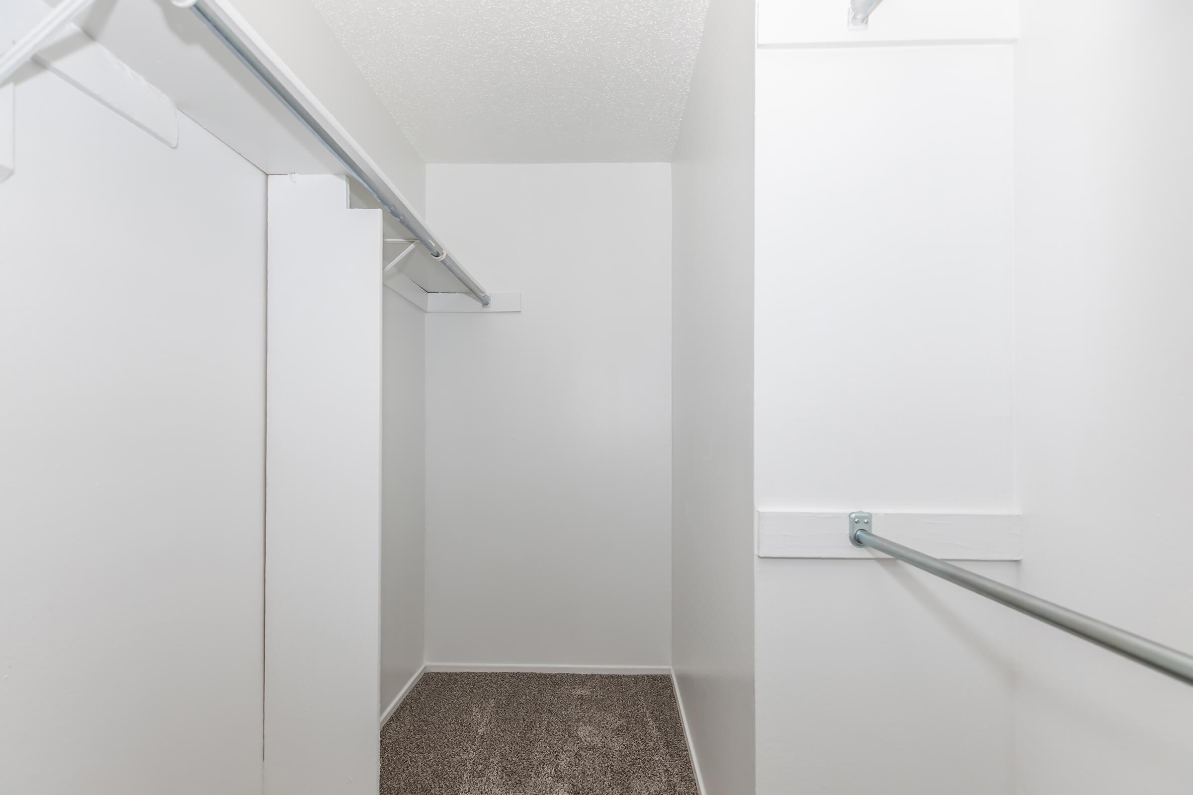A spacious, empty closet featuring white walls and a carpeted floor. Two hanging rods are visible on the left side, with ample room for storage. The overall space appears clean and well-maintained.