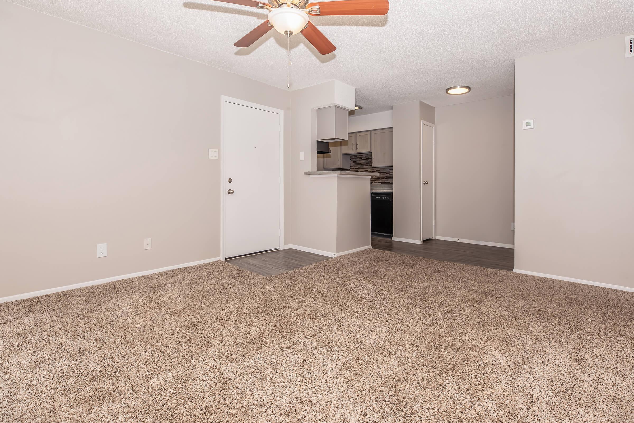 An interior view of an apartment featuring a spacious carpeted living area with a ceiling fan. There's a door leading outside and an open kitchen area in the background with a breakfast bar. The walls are painted light beige, and the flooring is a combination of carpet and hardwood.