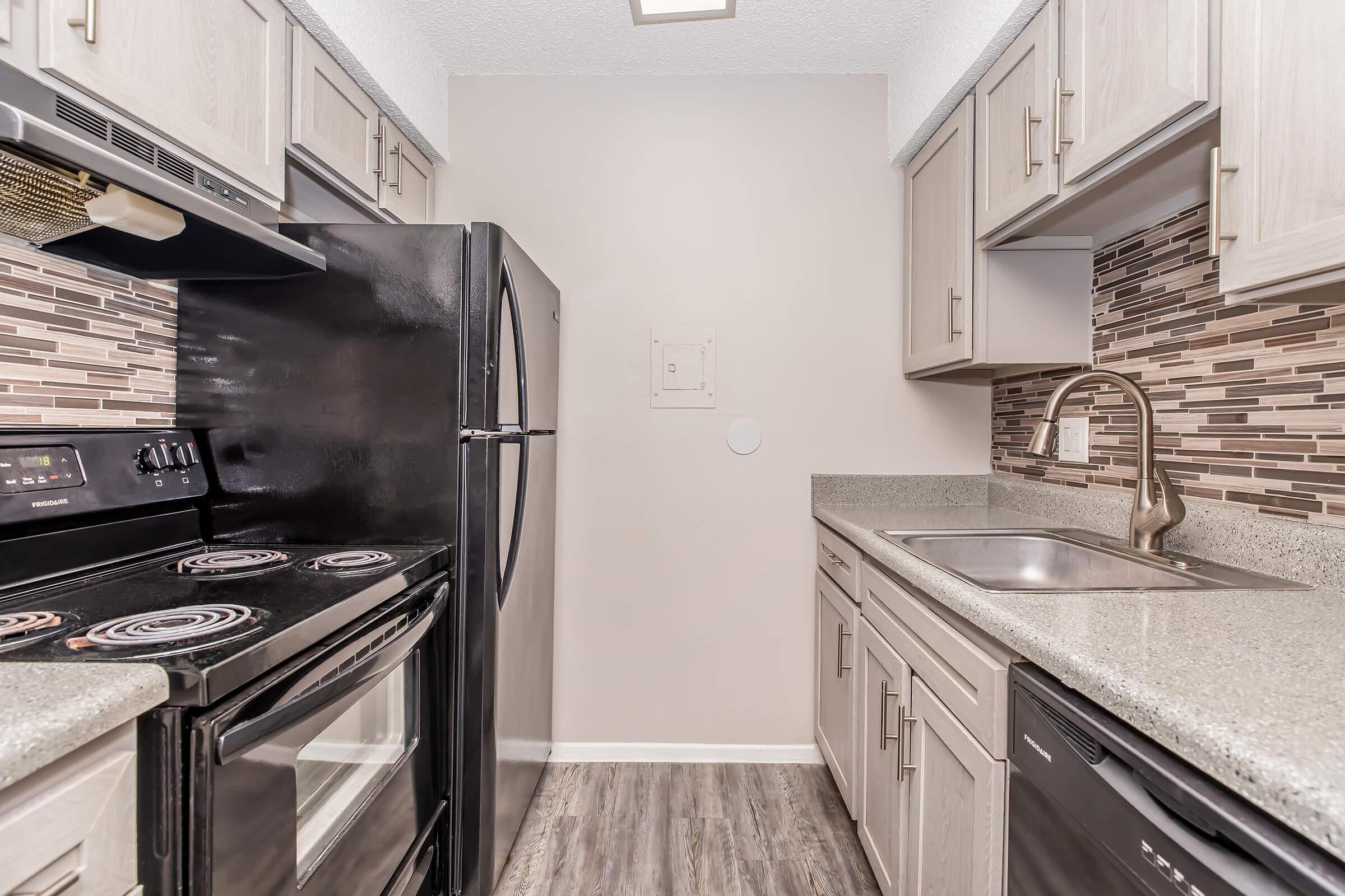 Modern kitchen featuring dark appliances, including a stove and refrigerator, with light-colored cabinets and countertops. The backsplash consists of small tiles, adding texture. The space is well-lit, creating a welcoming atmosphere. The layout is compact and functional, ideal for cooking and meal preparation.