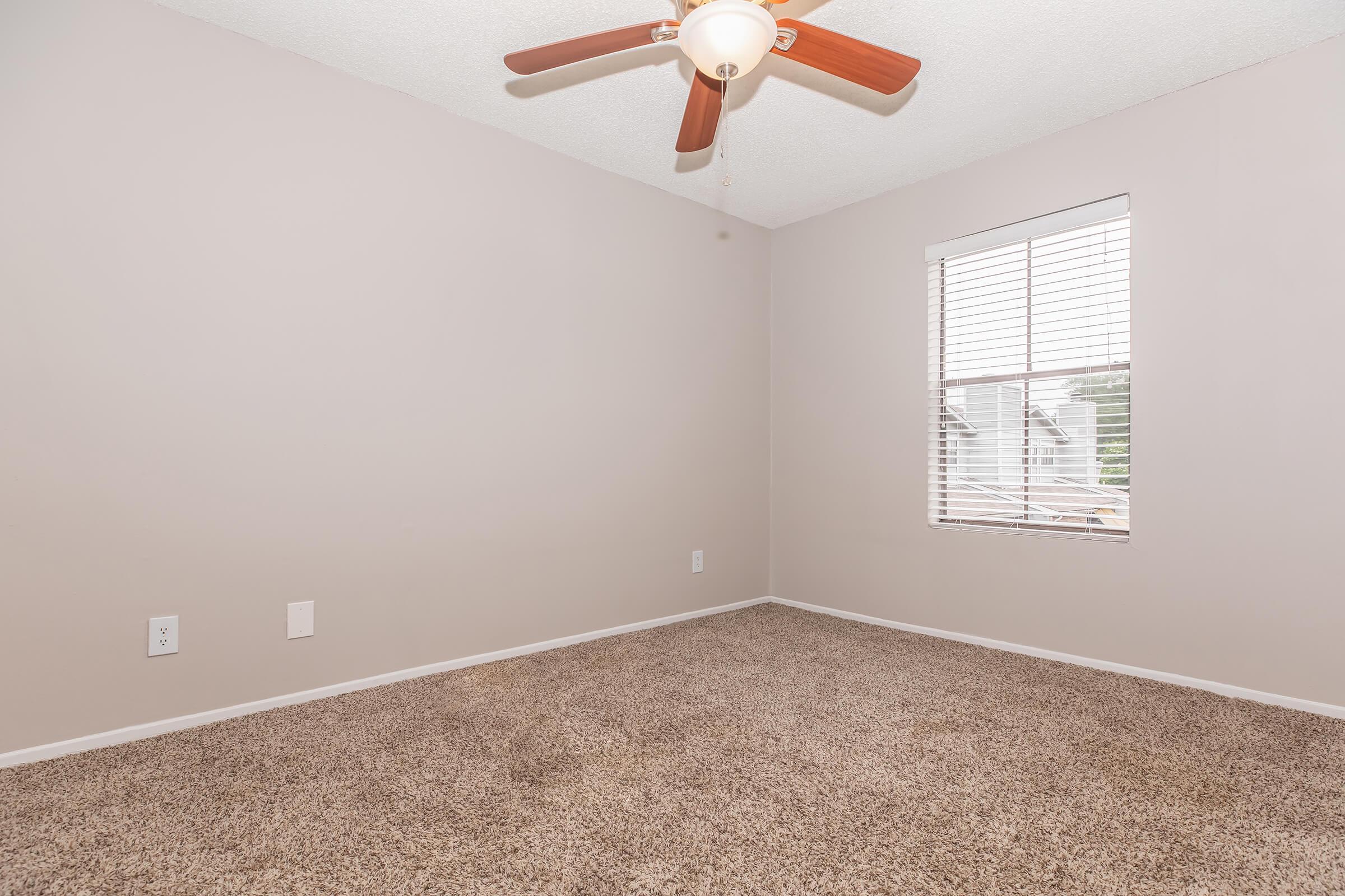 A well-lit empty room with light beige walls and a ceiling fan. The floor is covered with carpet, and there is a window with blinds letting in natural light. The room is spacious and uncluttered, suitable for various purposes like a bedroom or office.