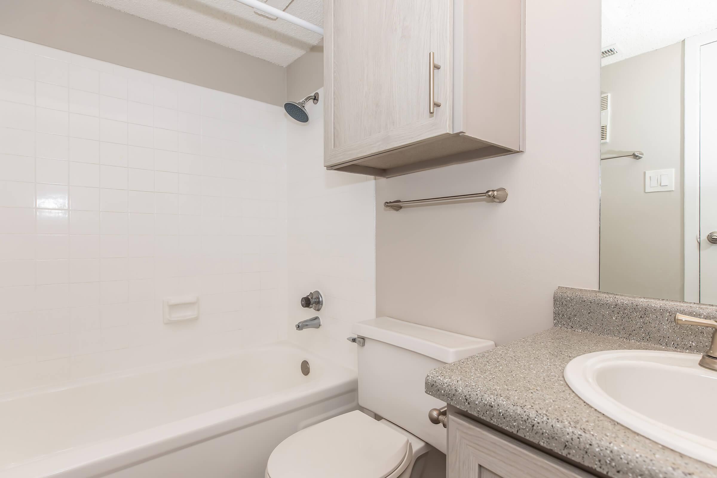 A clean and modern bathroom featuring a white tiled shower and bathtub combination, a single sink with a granite countertop, a mirrored cabinet above the sink, and chrome fixtures. The walls are painted in a light, neutral color, creating a bright and airy atmosphere.