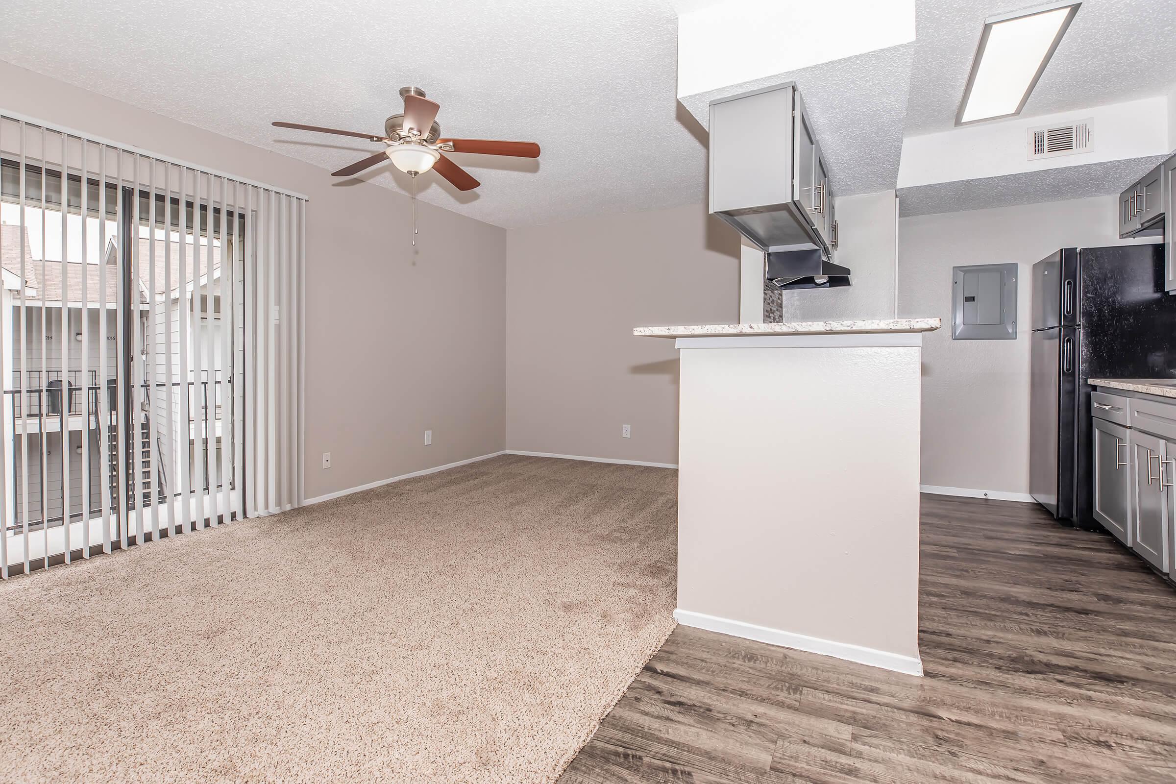 A spacious living area with light-colored walls and a ceiling fan. There's a window with vertical blinds and a sliding door leading to a balcony. The flooring features carpet in the living space and a hardwood-style finish in the kitchen area, which has modern appliances and a breakfast bar.