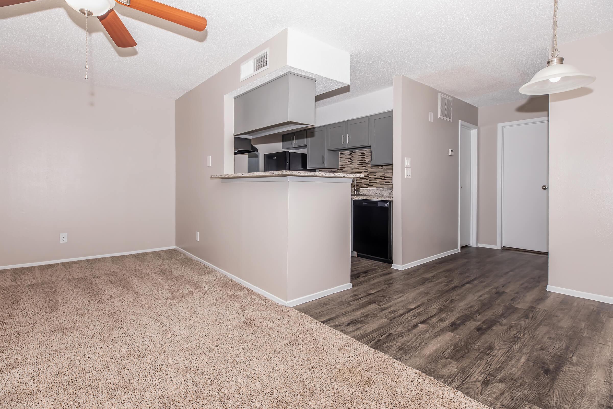 Interior view of an apartment featuring a spacious living area with carpeted flooring, a ceiling fan, and an open layout leading to a modern kitchen with gray cabinets and a dark countertop. There’s a doorway leading to another room and a light fixture hanging in the dining area. Bright, neutral wall colors create a welcoming atmosphere.