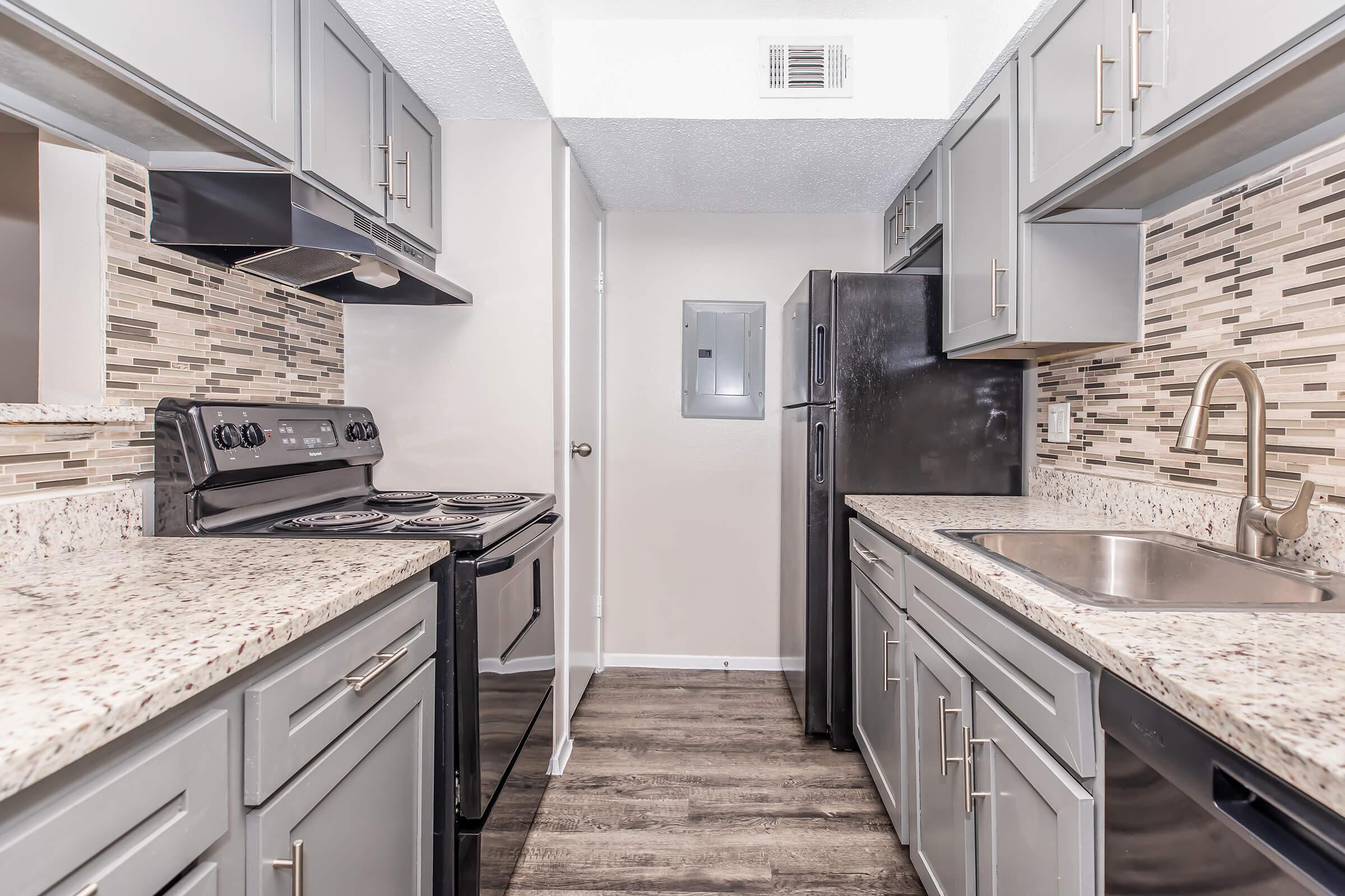 Modern kitchen featuring gray cabinetry, granite countertops, a black refrigerator, and an electric stove. The backsplash consists of mosaic tiles, and there is a stainless steel sink with a chrome faucet. The space is well-lit with neutral walls and floor tiles, creating a contemporary aesthetic.